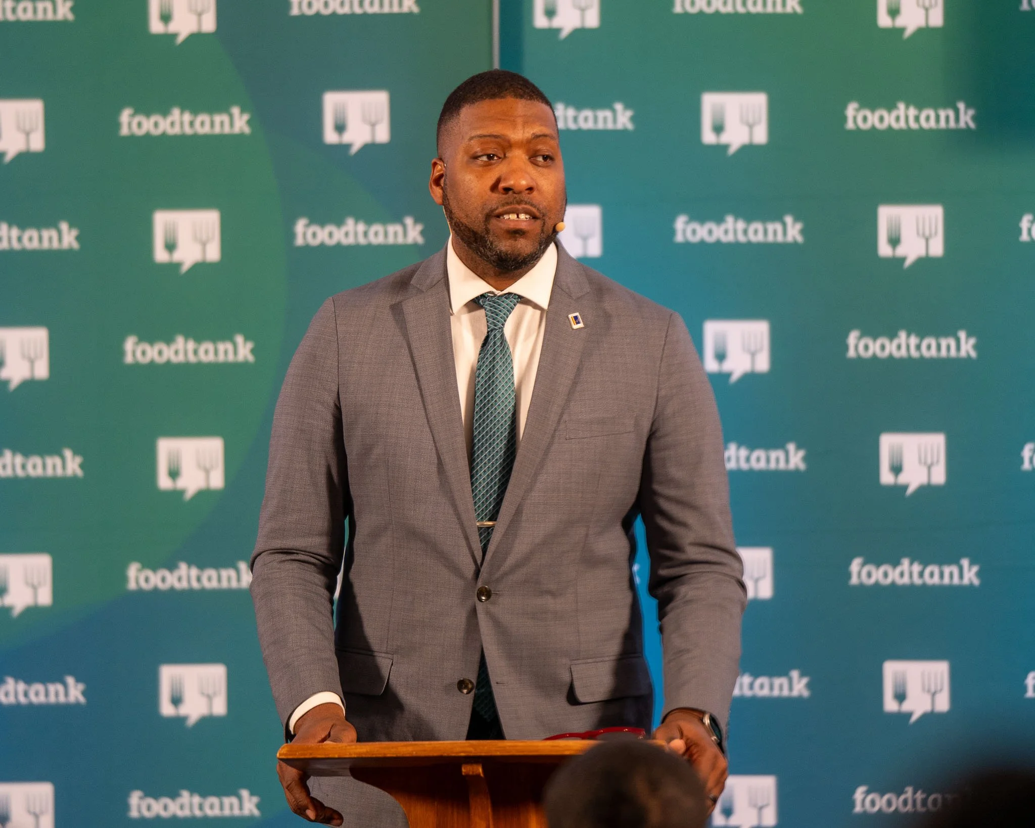 A man in a suit and tie speaks at a podium with a "foodtank" logo backdrop.