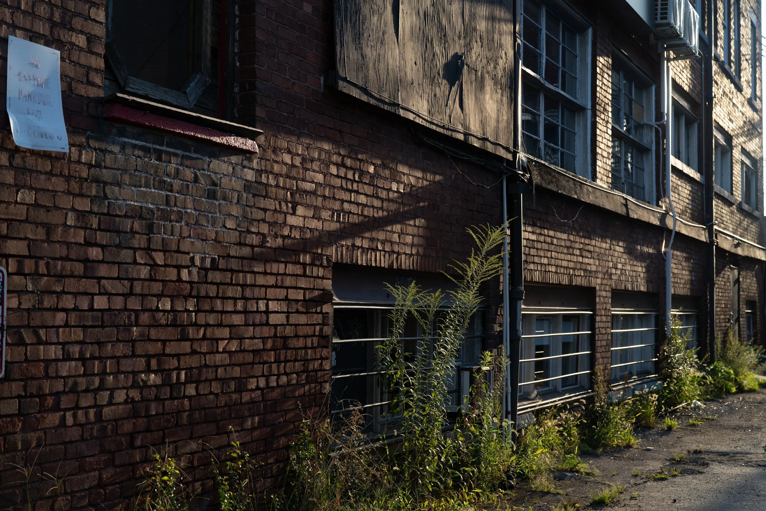 Side view of an old brick building with boarded-up windows, overgrown weeds, and weathered facade.