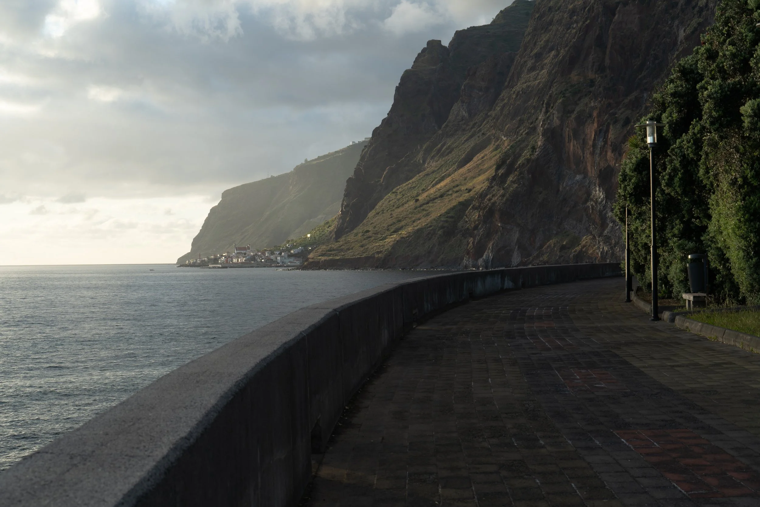 Coastal walkway with ocean view on the left and steep cliffs on the right, lined with trees and street lamps under a cloudy sky.