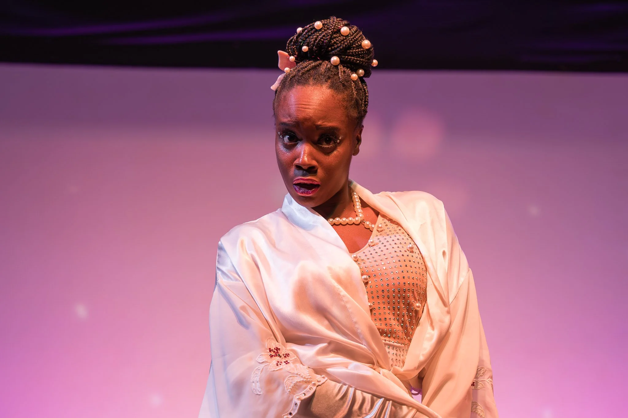 A woman in a white satin robe and pearl necklace, with braided hair adorned with pearls, making a dramatic facial expression in front of a purple-lit background.