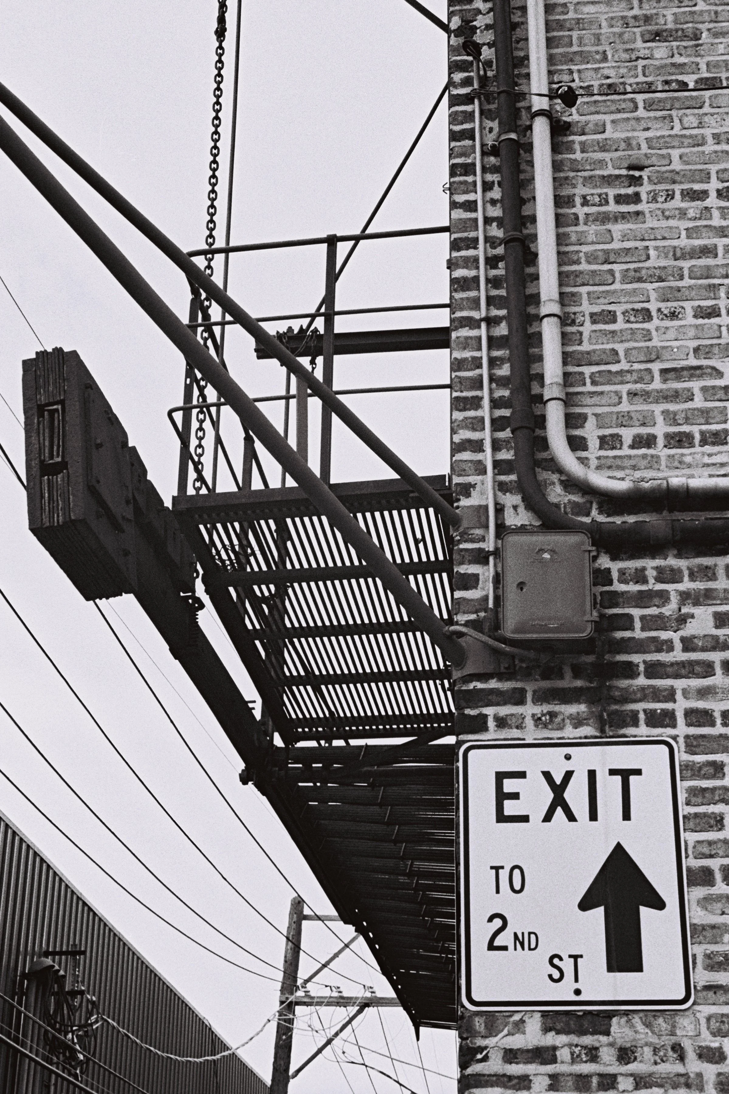 Black and white image of a brick building corner with a metal fire escape. An "EXIT to 2nd ST" sign points upward. Power lines run alongside the building.