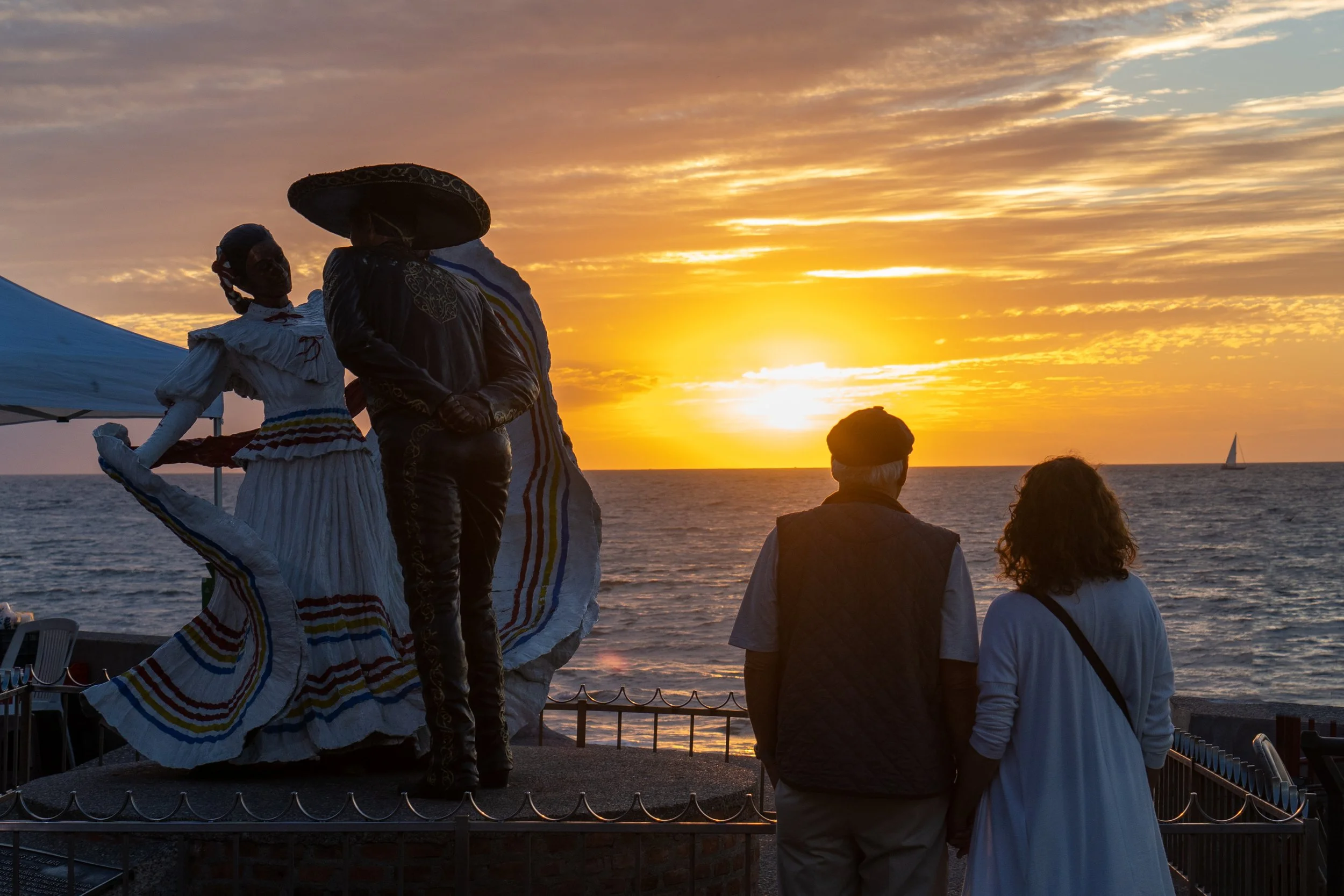 Two people watching a beach sunset near sculptures of a traditional Mexican couple, with a sailboat on the horizon.