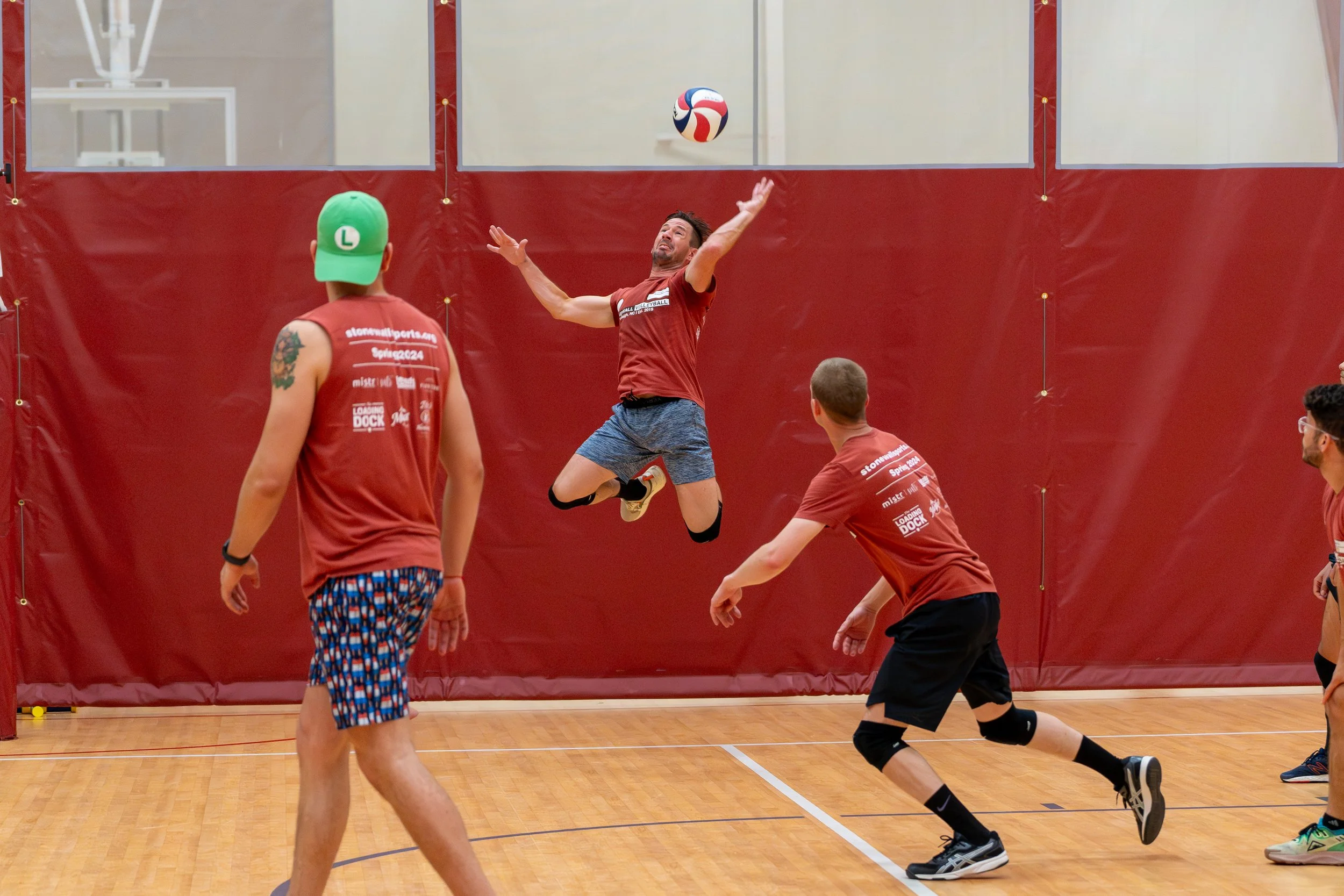 Indoor volleyball game with people in red shirts, one player jumping to hit the ball.