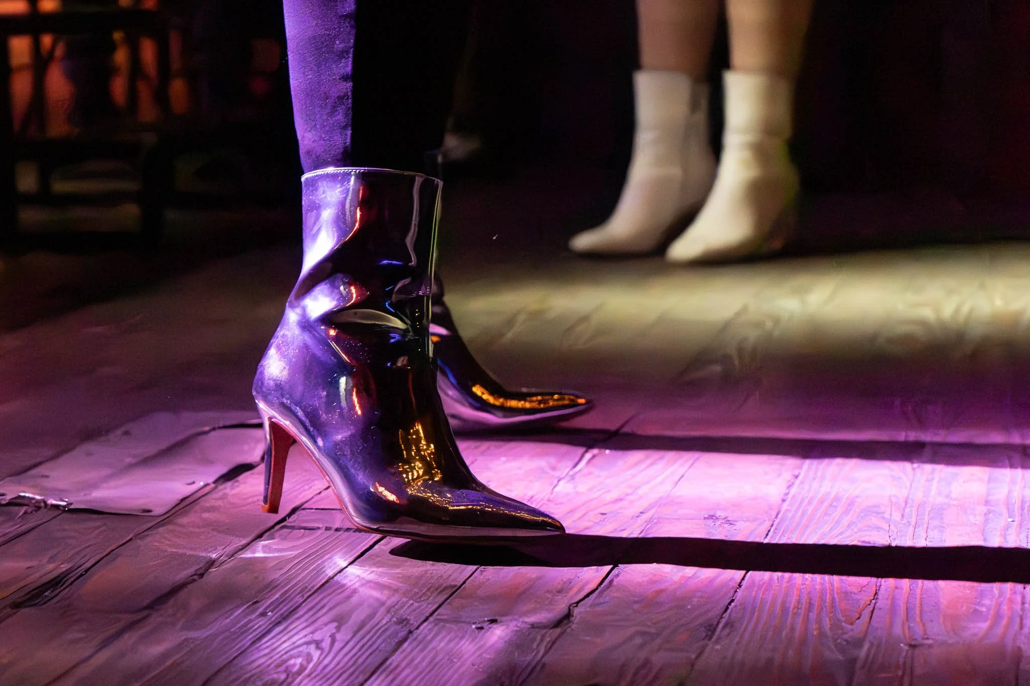 Women wearing shiny black ankle boots and white boots on a wooden floor with stage lighting.