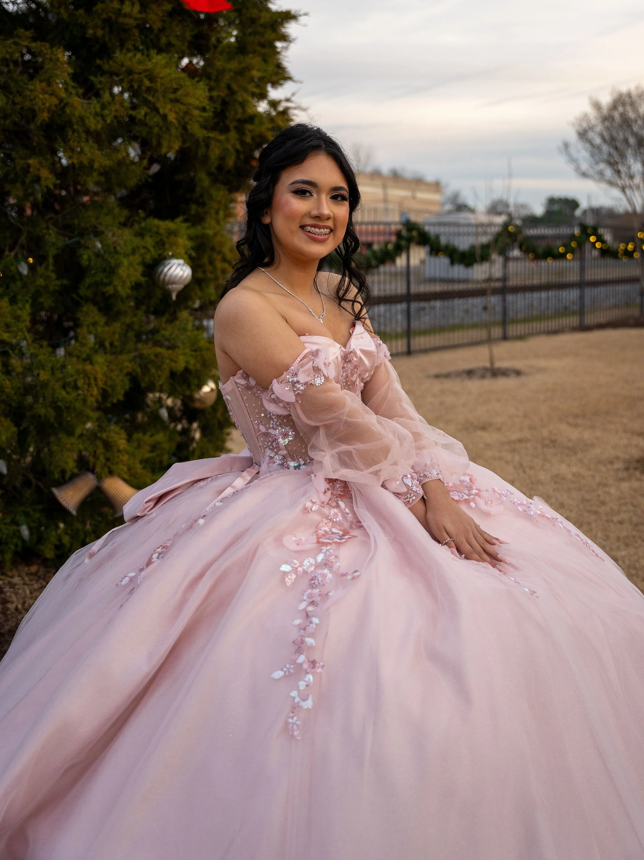 A young woman wearing a pink ball gown with floral embellishments and mesh sleeves is posing outdoors. She is smiling with braces and has dark wavy hair. A decorated tree with ornaments is in the background along with outdoor fencing and lights.