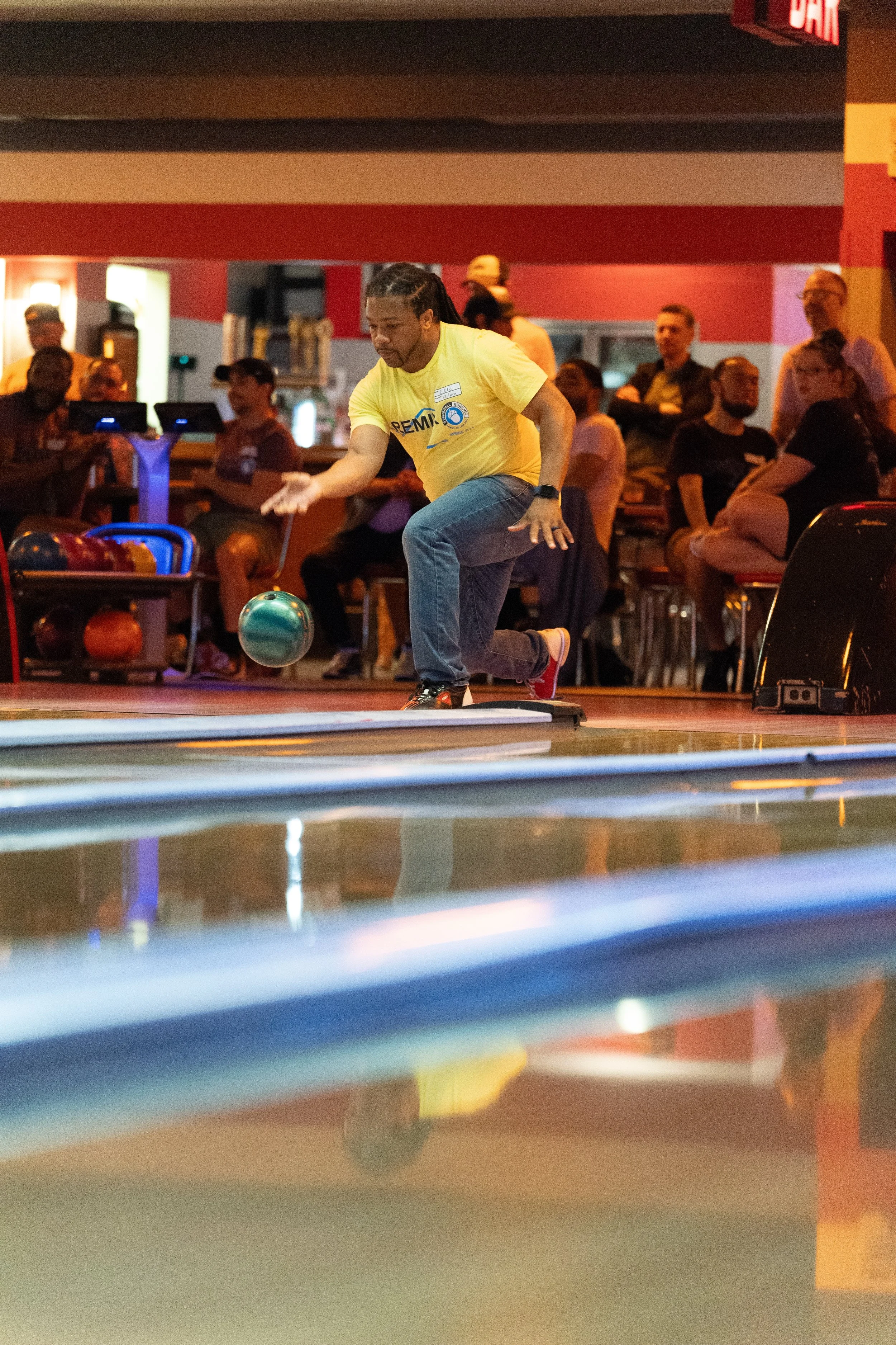 A man bowling in a bright, colorful bowling alley, surrounded by onlookers seated and standing near the lanes.
