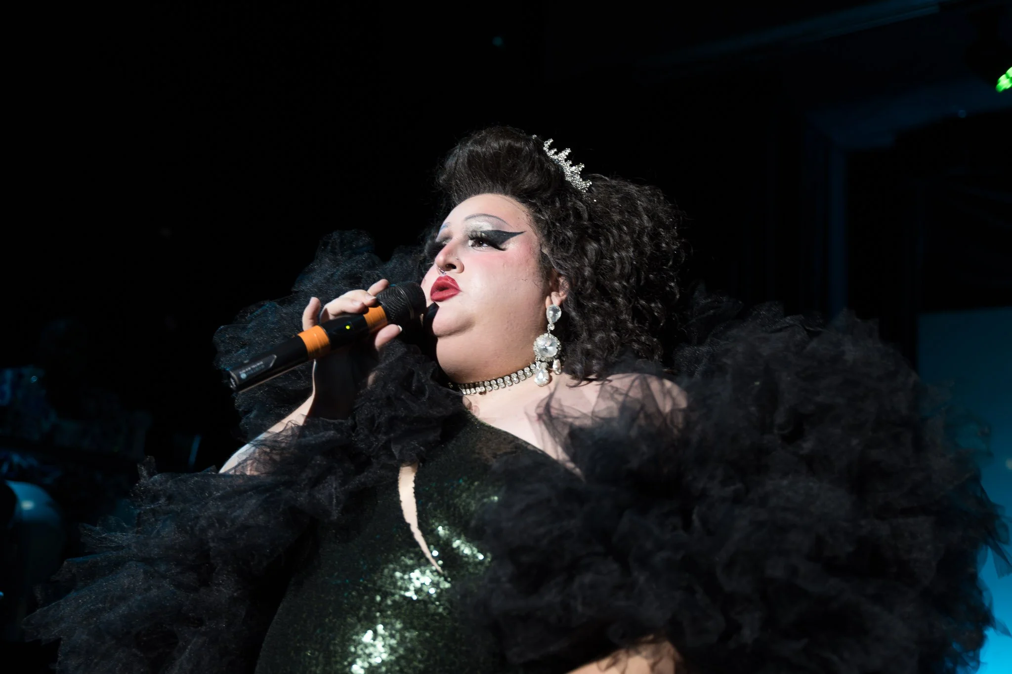 Drag performer in a glamorous black outfit singing on stage with dramatic makeup and accessories.