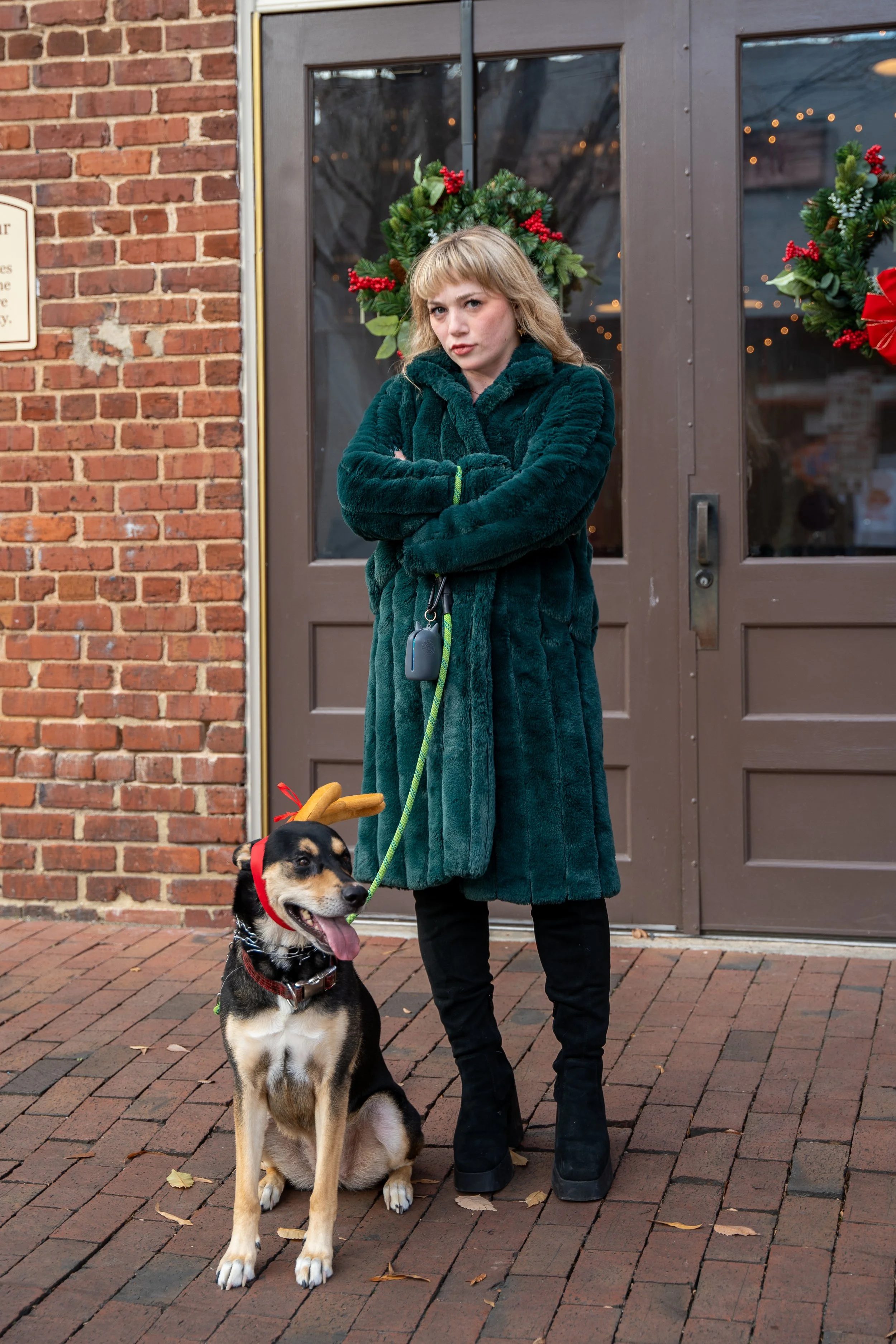 Woman in green fur coat standing with a dog wearing reindeer antlers on a leash, in front of a brick wall with holiday wreaths.