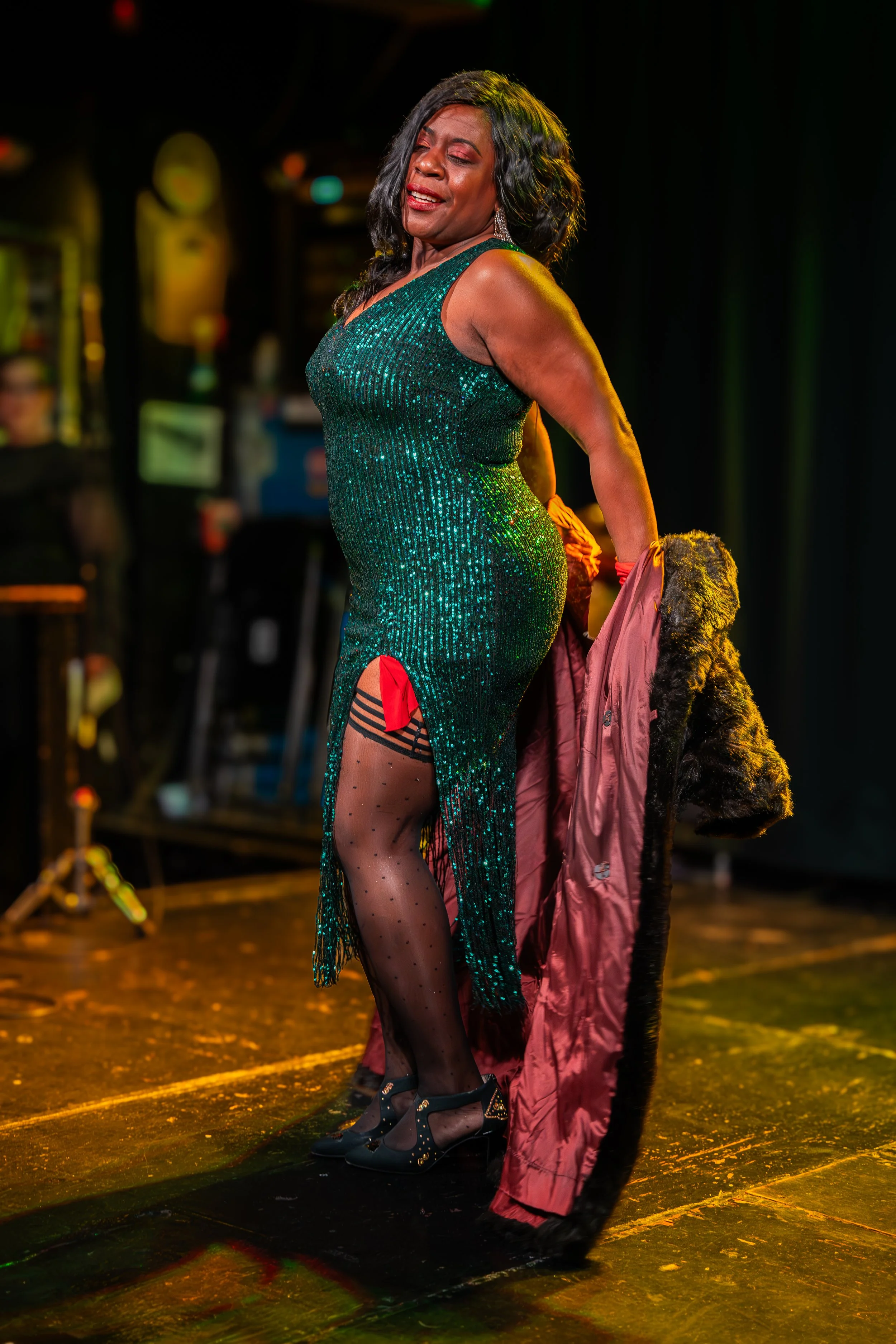A woman in a sequined green dress performing on stage, holding a coat, with dramatic lighting.