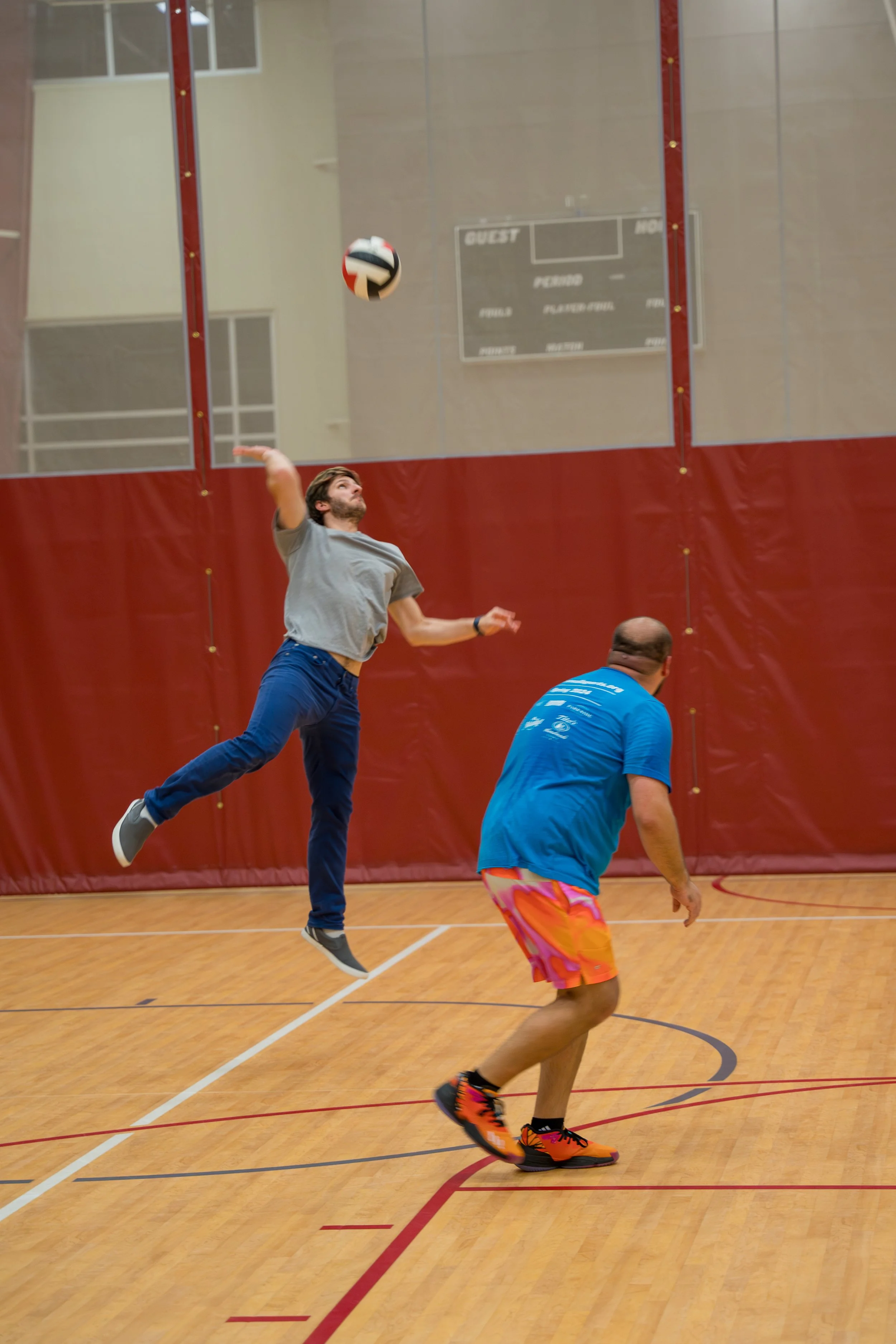 Two people playing volleyball in a gym; one is jumping to hit the ball.
