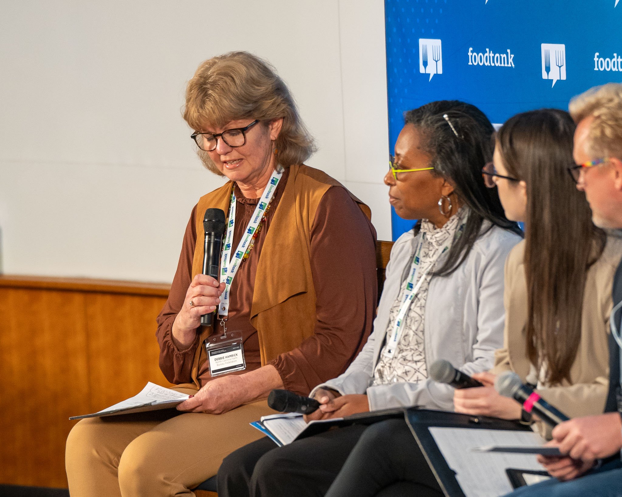 Panel discussion at Food Tank event with diverse group of speakers holding microphones and notes, seated in front of a branded backdrop.