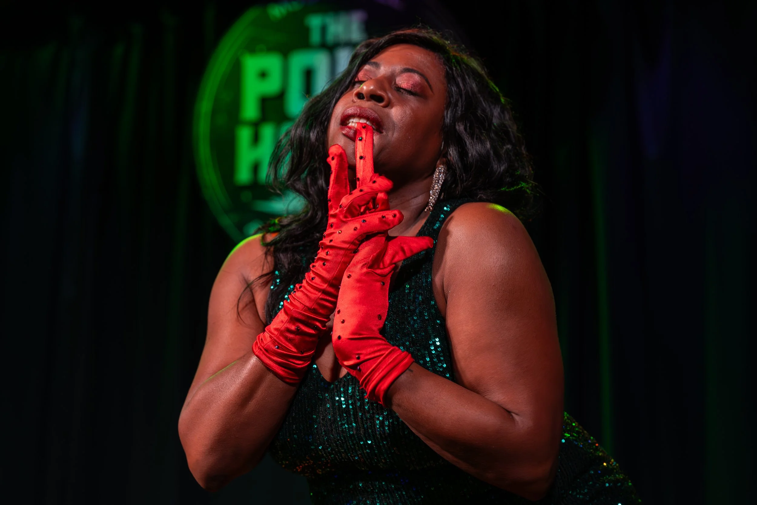 Person wearing red gloves and a sparkly dress, posing with fingers near face, in front of a green neon sign.