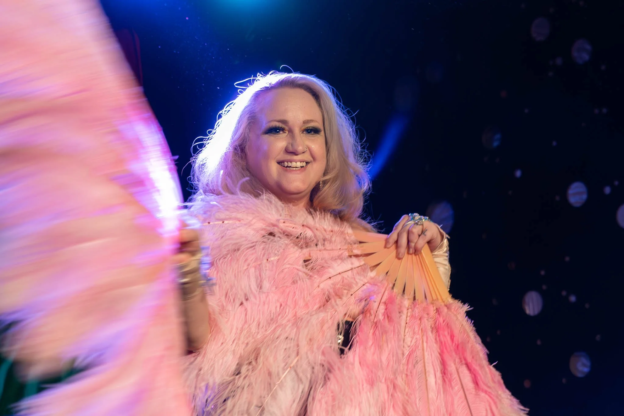 Person holding fluffy pink feather fans on stage with colorful lighting.