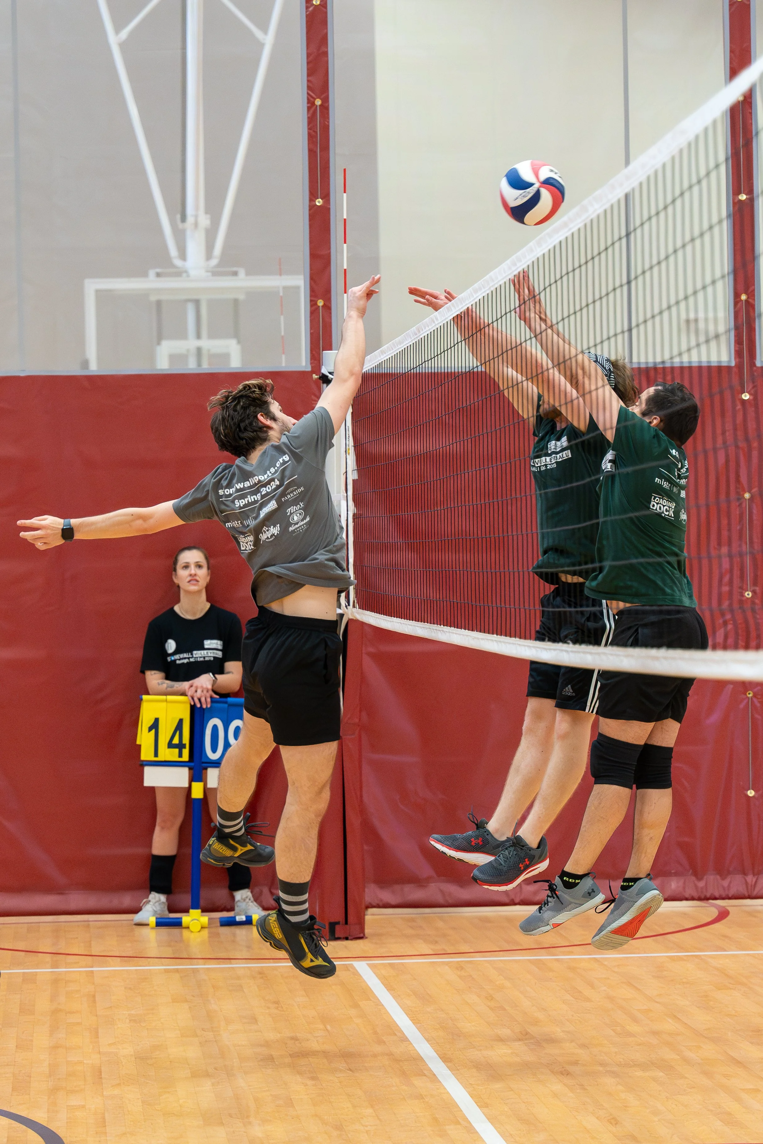 Three volleyball players in action at the net during an indoor game, with one player attempting a spike and two players blocking. A referee is in the background with a scoreboard showing the score "14-09." The players are wearing sports jerseys and s