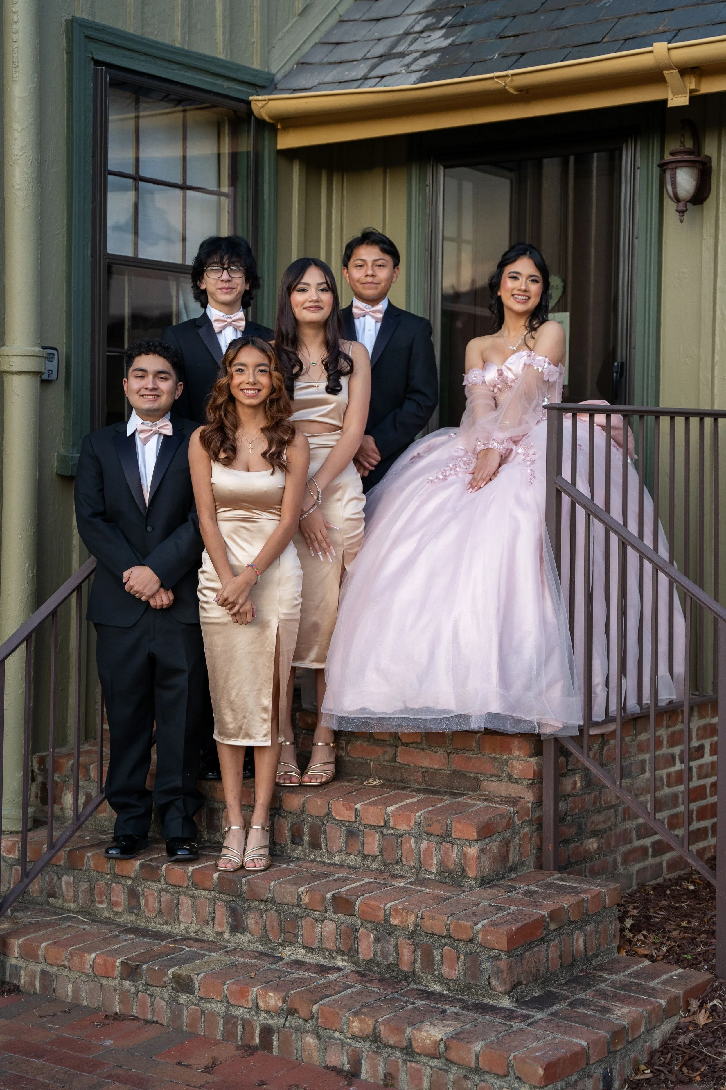 Group of young people in formal attire on a brick staircase, with one wearing a pink ball gown.