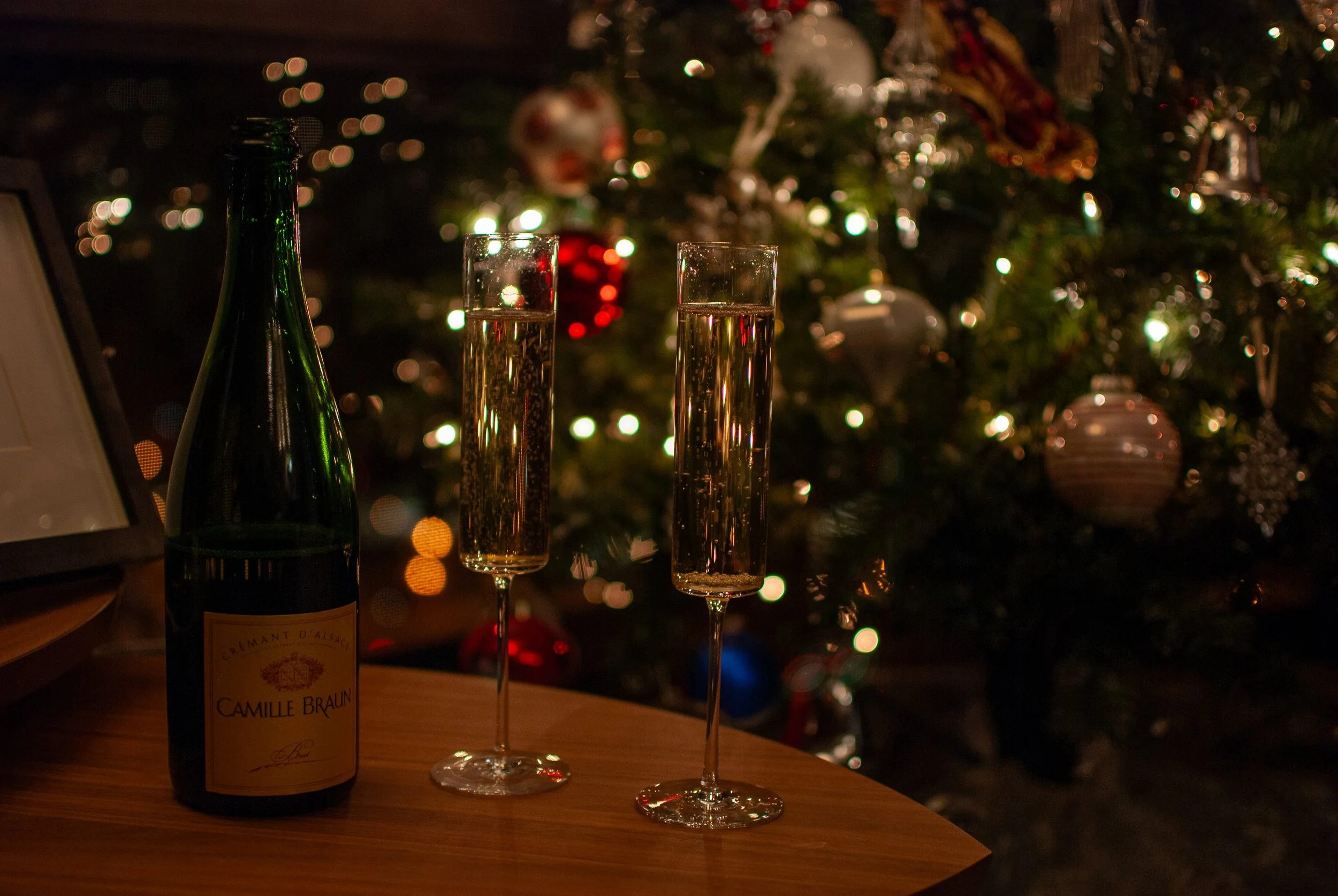Bottle and two flutes of champagne on a table with a decorated Christmas tree in the background.