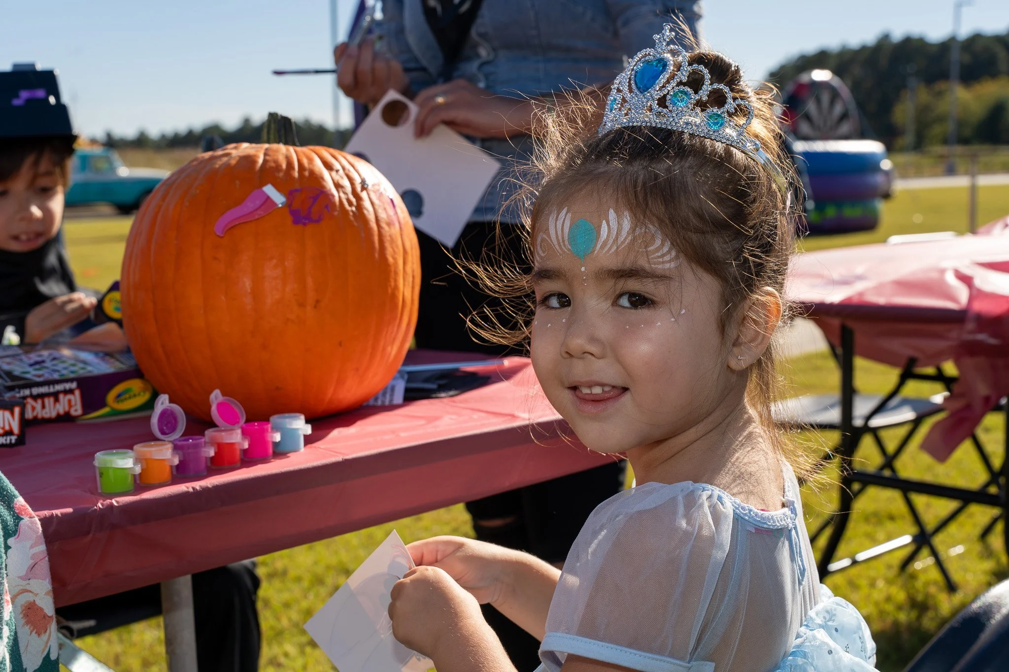 Young girl wearing a jeweled tiara and face paint at a pumpkin painting activity outdoors