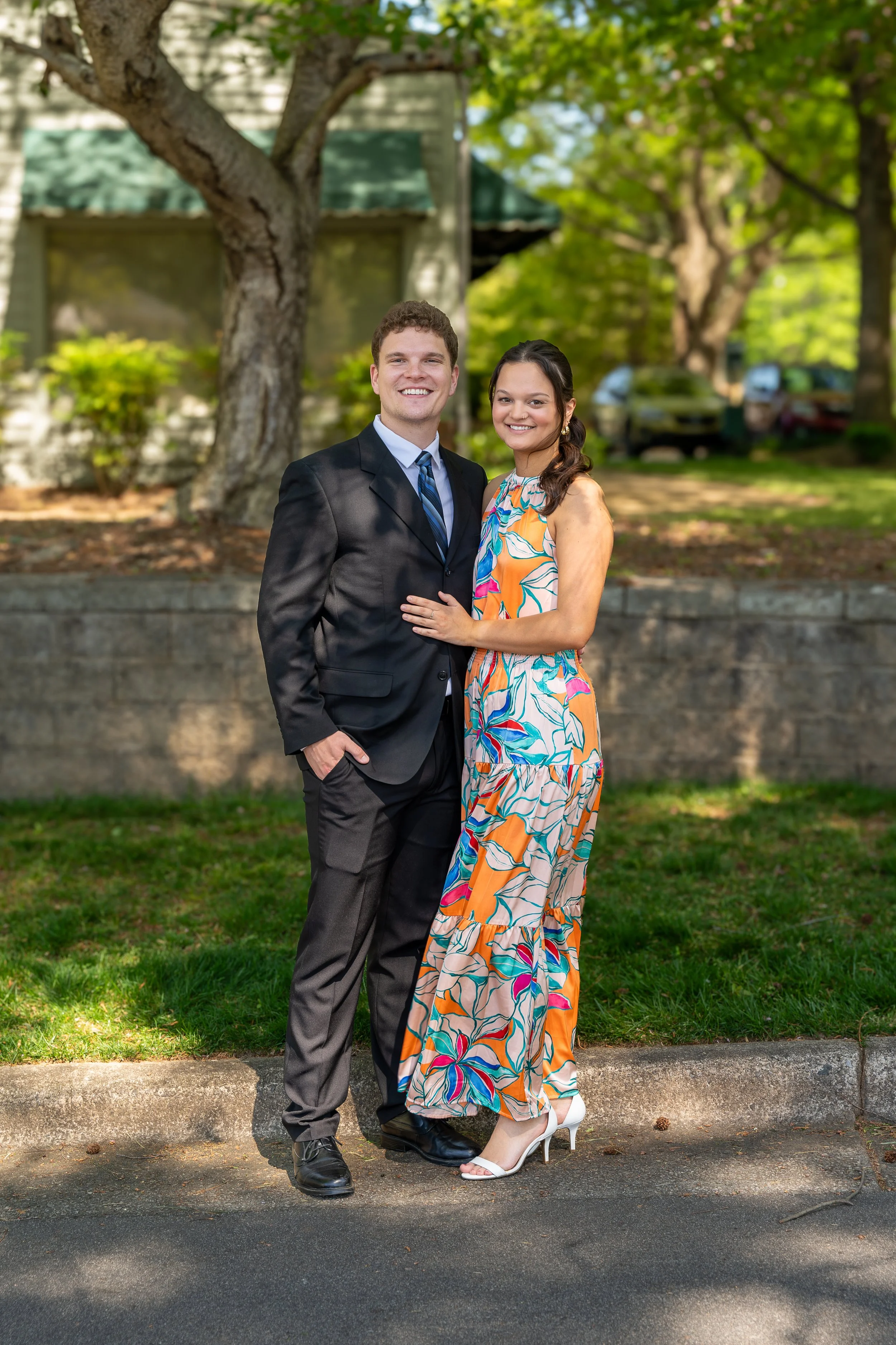 A couple posing outdoors, the man in a black suit and tie, and the woman in a colorful floral dress. They are standing on a path with trees and a building in the background.