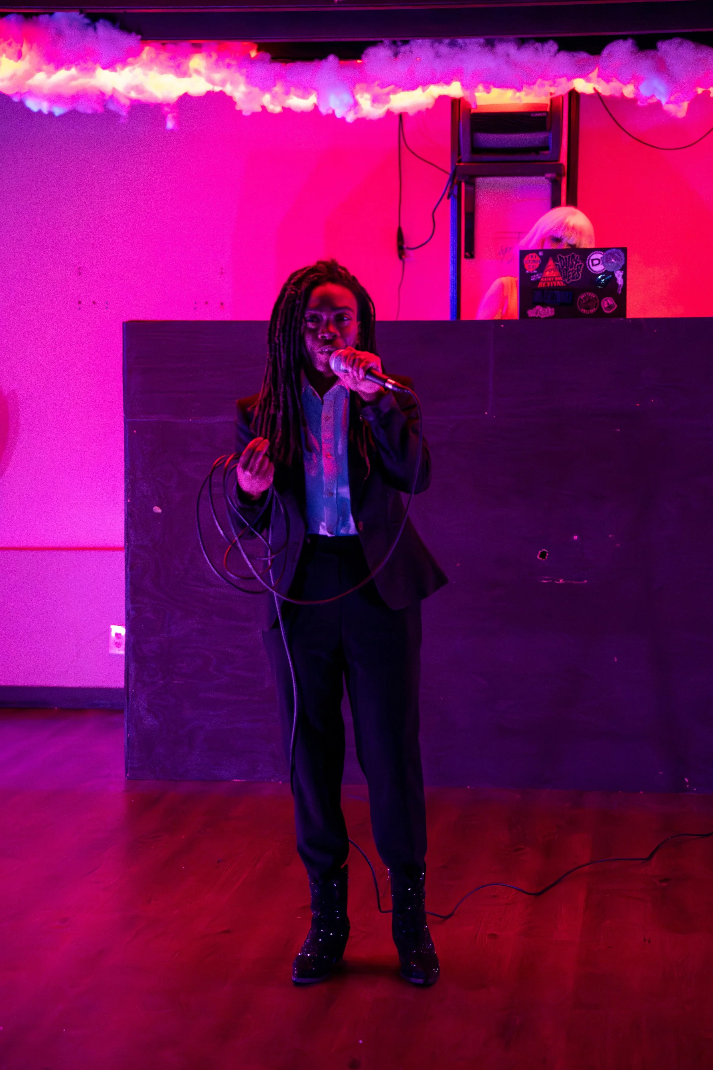 Person performing with a microphone on stage under purple and pink lighting, DJ visible in background with a laptop.