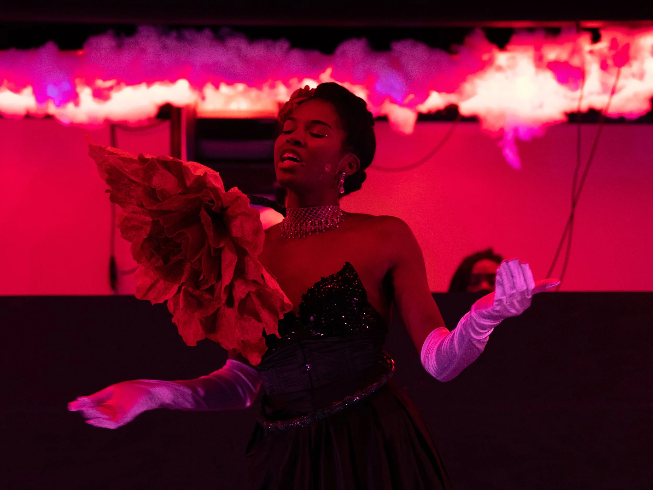 Performer in glamorous attire, wearing long white gloves and holding a large flower-like prop, sings under red stage lighting.