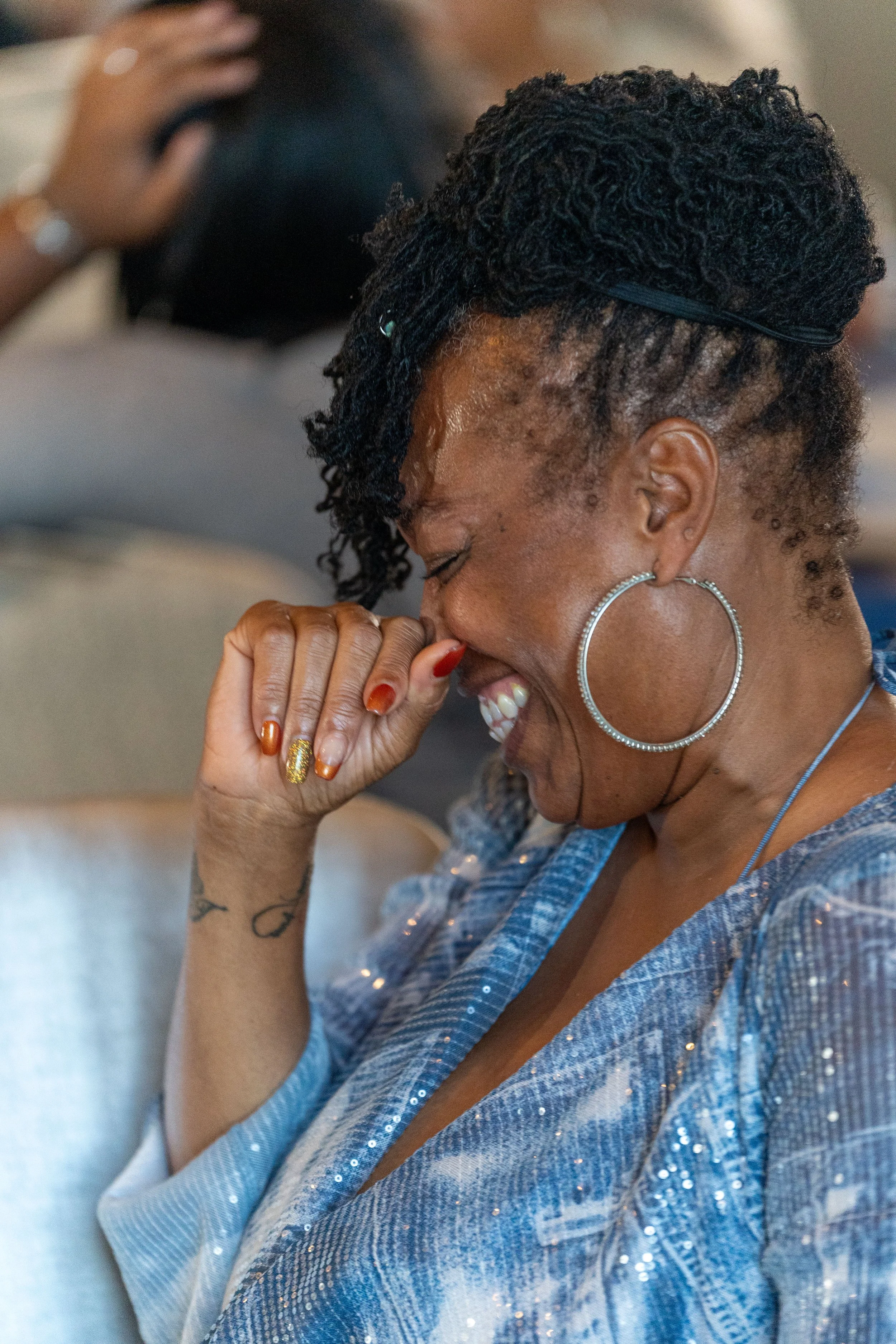 Woman smiling and covering her mouth, wearing a blue sequined top and large hoop earrings, with styled hair and colorful nails.