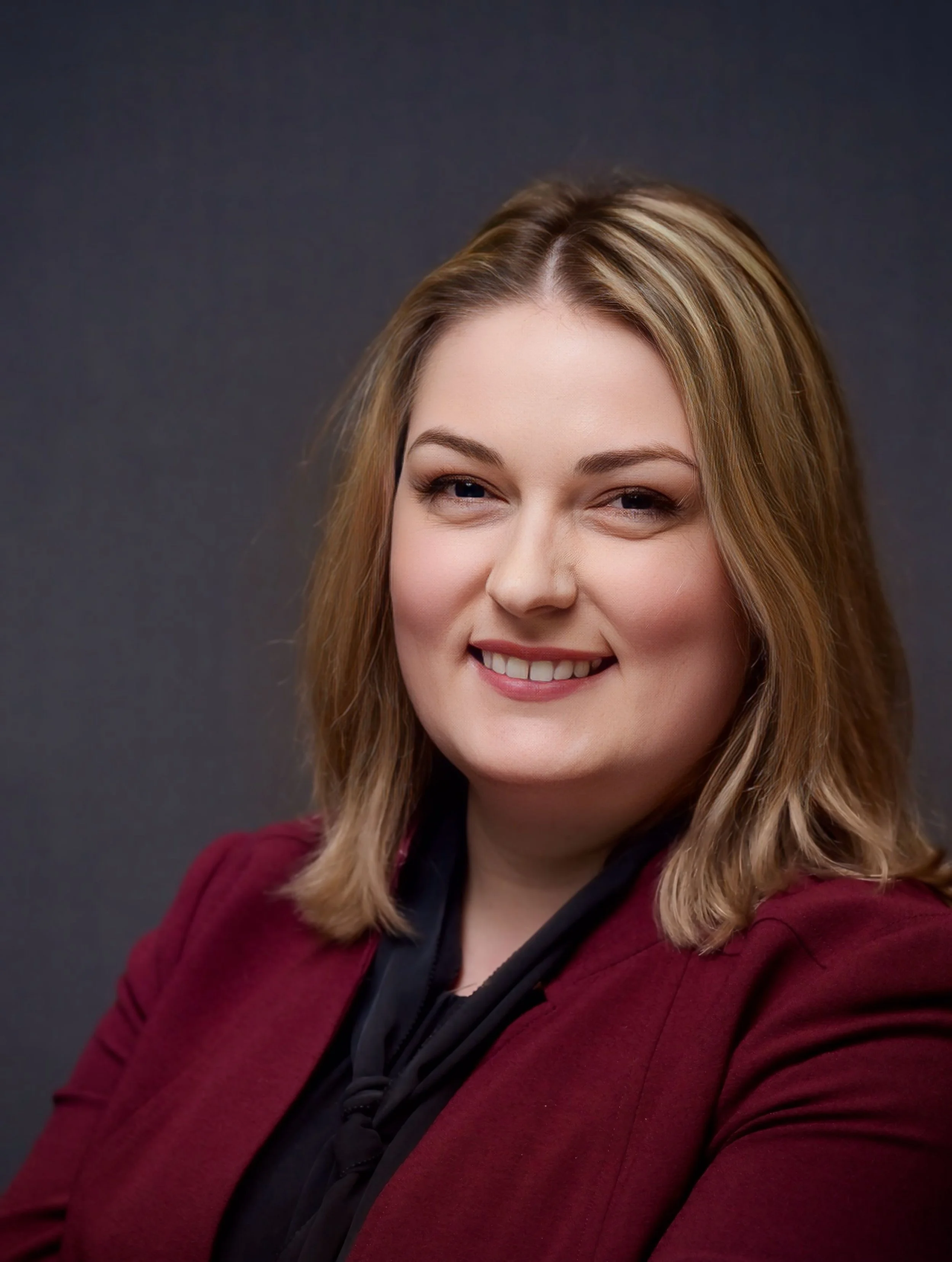 A woman with blonde hair styled with soft curls, wearing a maroon blazer and a black top, smiling against a dark gray background.