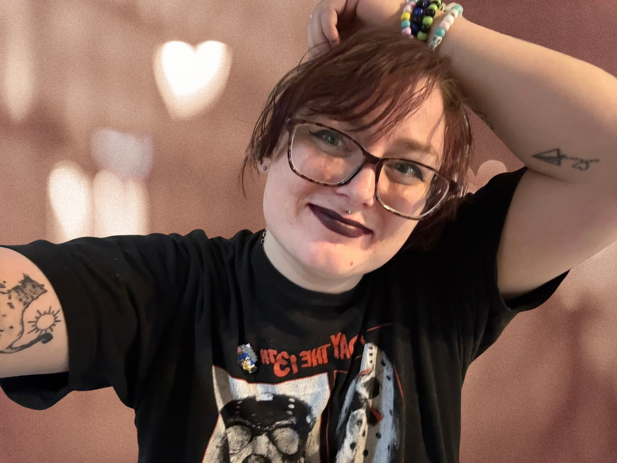 A person with short, reddish hair, wearing glasses, smiling, with tattoos on their left arm, and wearing colorful bracelets, posing in front of a pink background with heart-shaped bokeh lights.