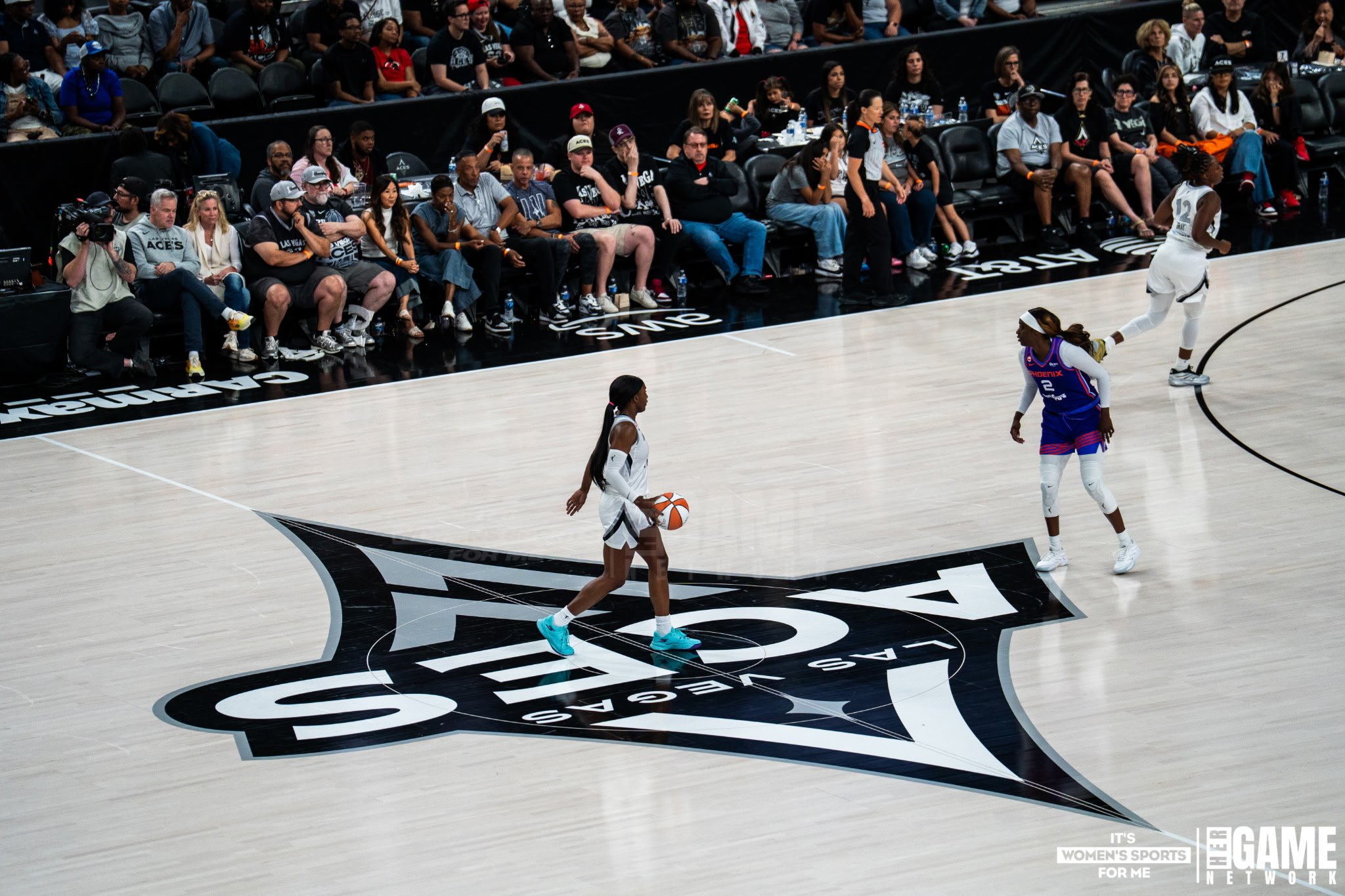 Women playing basketball during a game with spectators in the background.