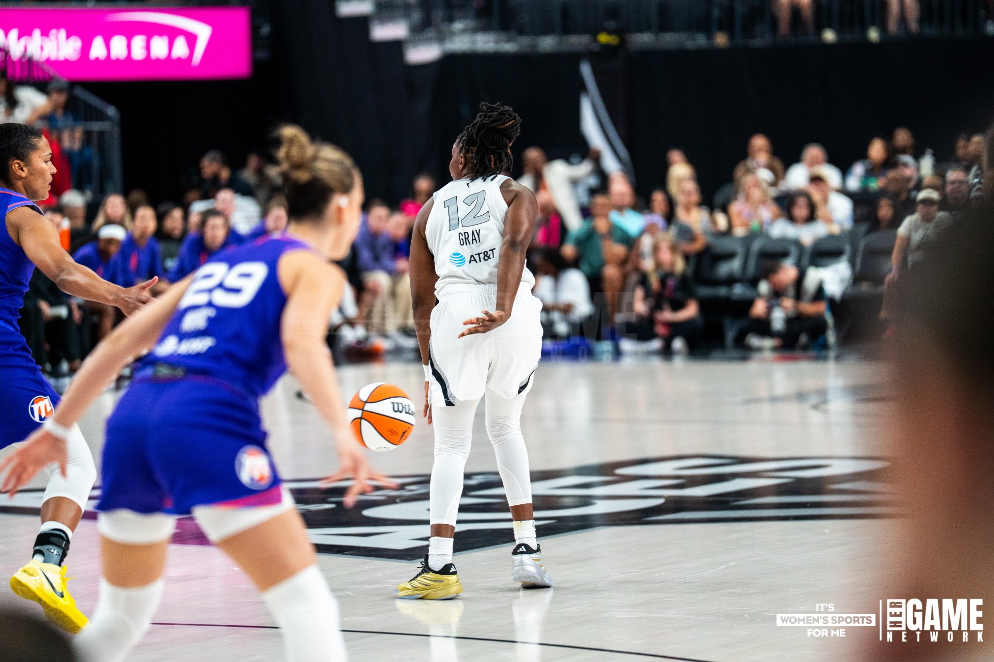 Women playing basketball in an indoor court, with one woman in white jersey holding a basketball, and others in blue uniforms in the background, spectators watching.
