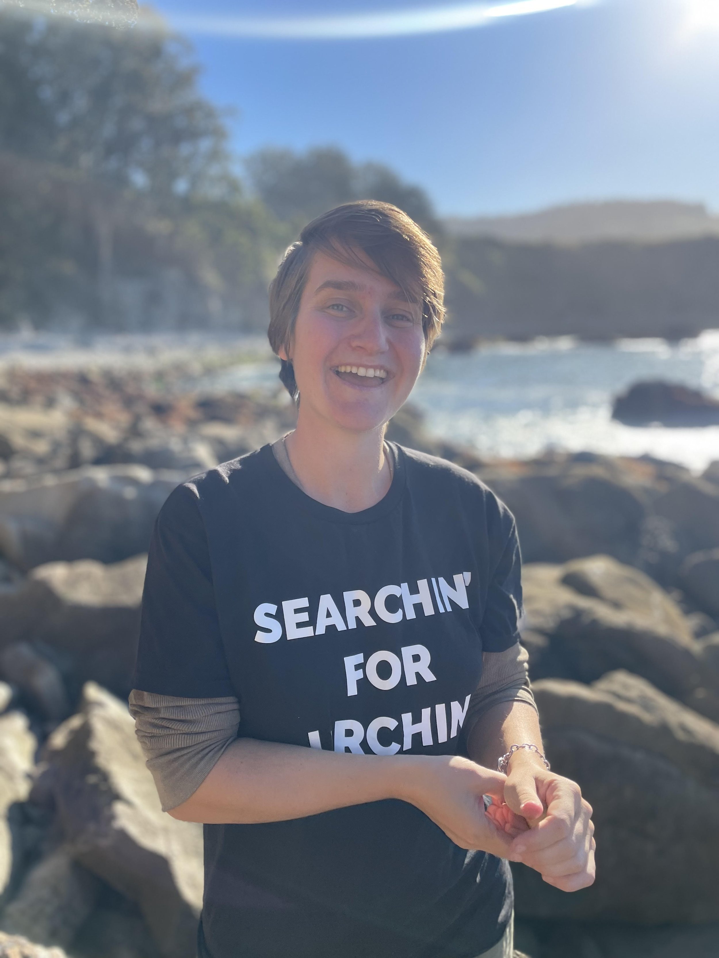 A smiling woman with short hair, wearing a black t-shirt with white text, stands outdoors near a rocky shoreline with water and hills in the background under a clear blue sky.