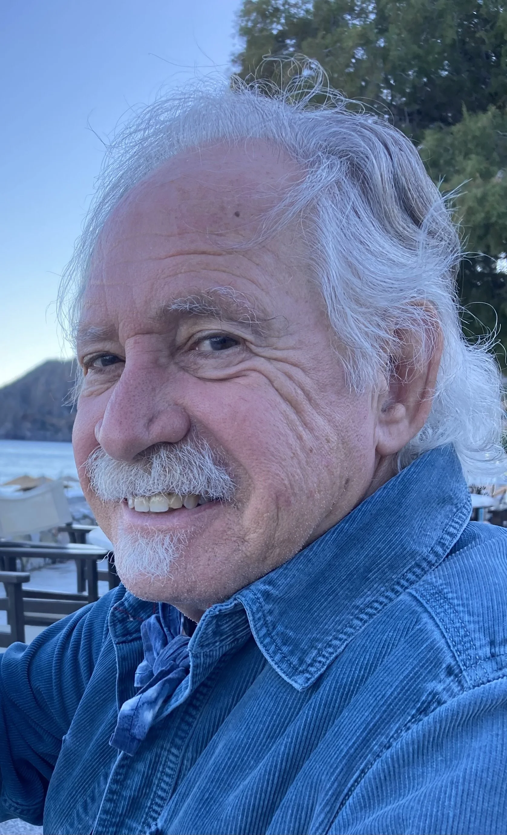 A smiling older man with white hair and a white mustache, wearing a blue striped shirt, outdoors near water with trees and a mountain in the background.