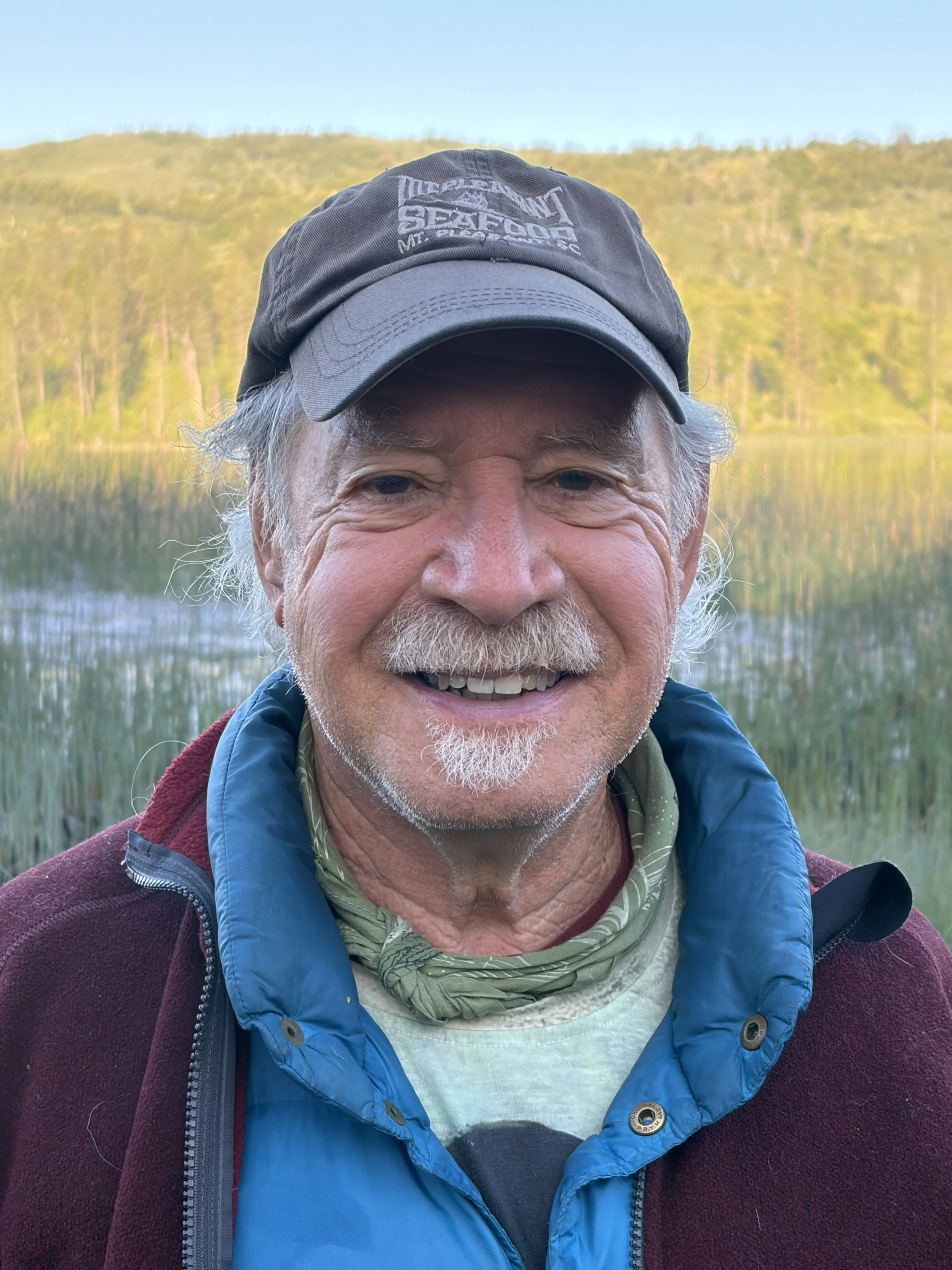 Close-up of an older man with gray hair and beard smiling outdoors, wearing a blue hoodie, maroon jacket, and a gray baseball cap, with a lake and hills in the background.