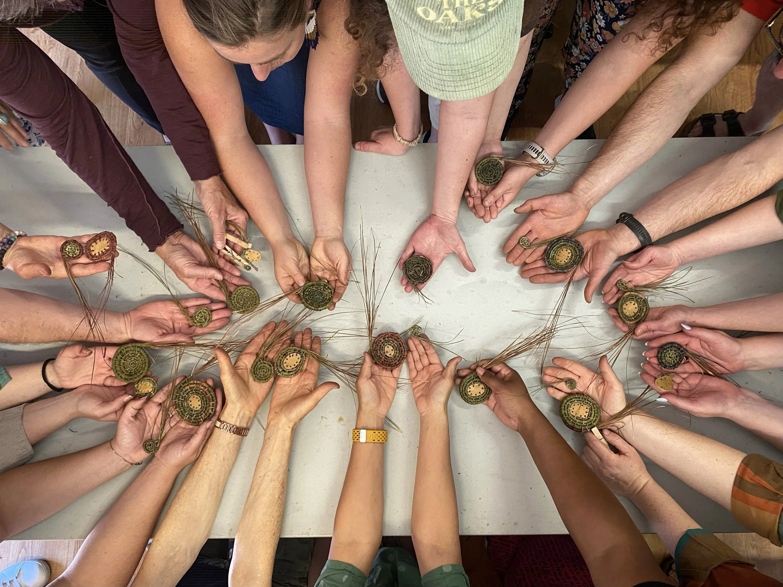 A top-down view of multiple hands reaching toward a table, holding woven circular objects, with some hands wearing watches, bracelets, and rings, indicating a group activity or craft session.
