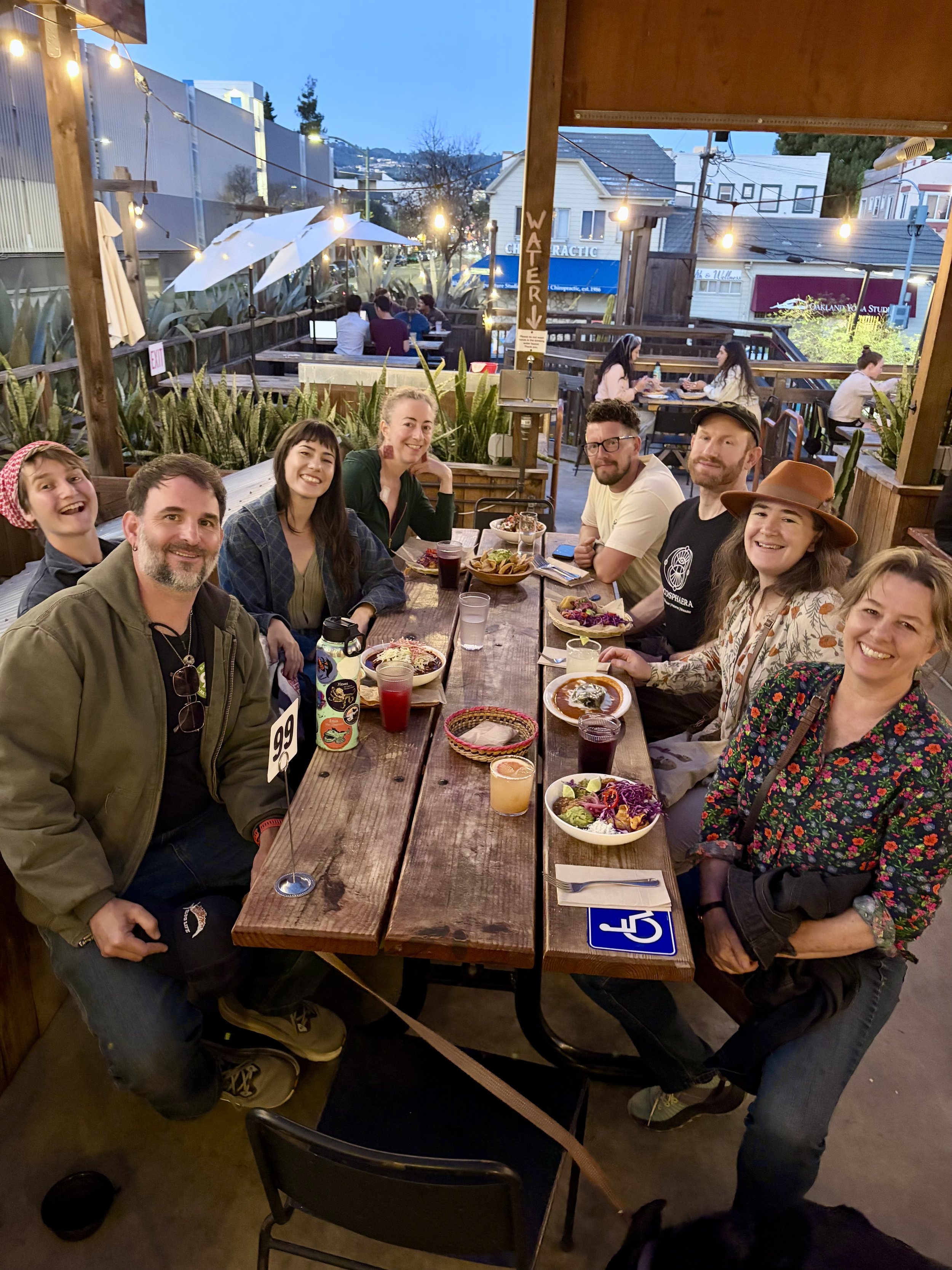 A group of seven people sitting at a wooden outdoor restaurant table during evening, smiling, with various dishes and drinks, some wearing casual and others wearing hats, with string lights and outdoor setting in the background.