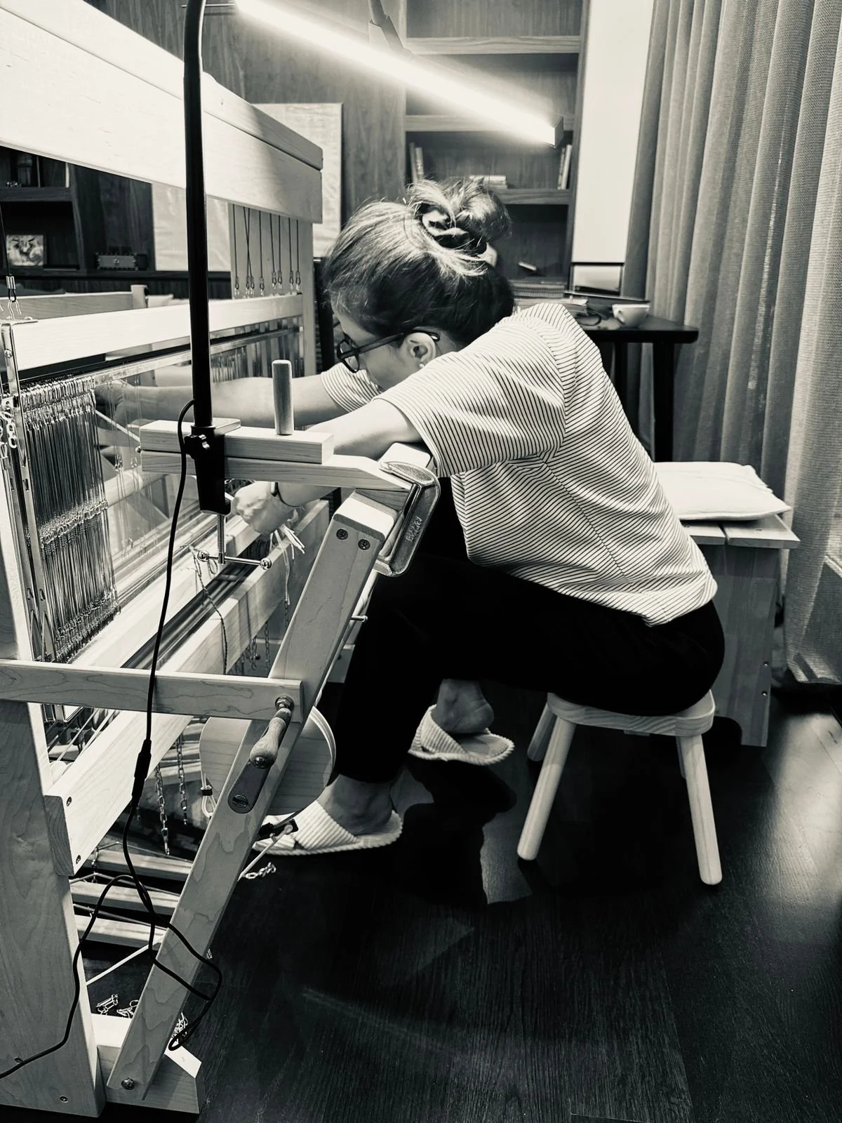 A woman is sitting on a stool, working on a knitting machine in a cozy, wood-paneled room. She has her hair tied up with a scrunchie, is wearing glasses, a striped shirt, black pants, and slippers.