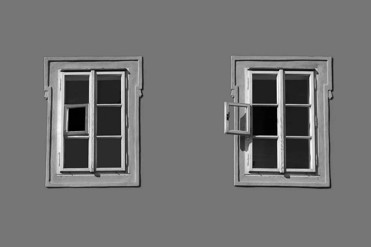 Two old wooden windows with shutters on a plain gray wall; one window has a small open window within it, and the other window's shutters are slightly open.