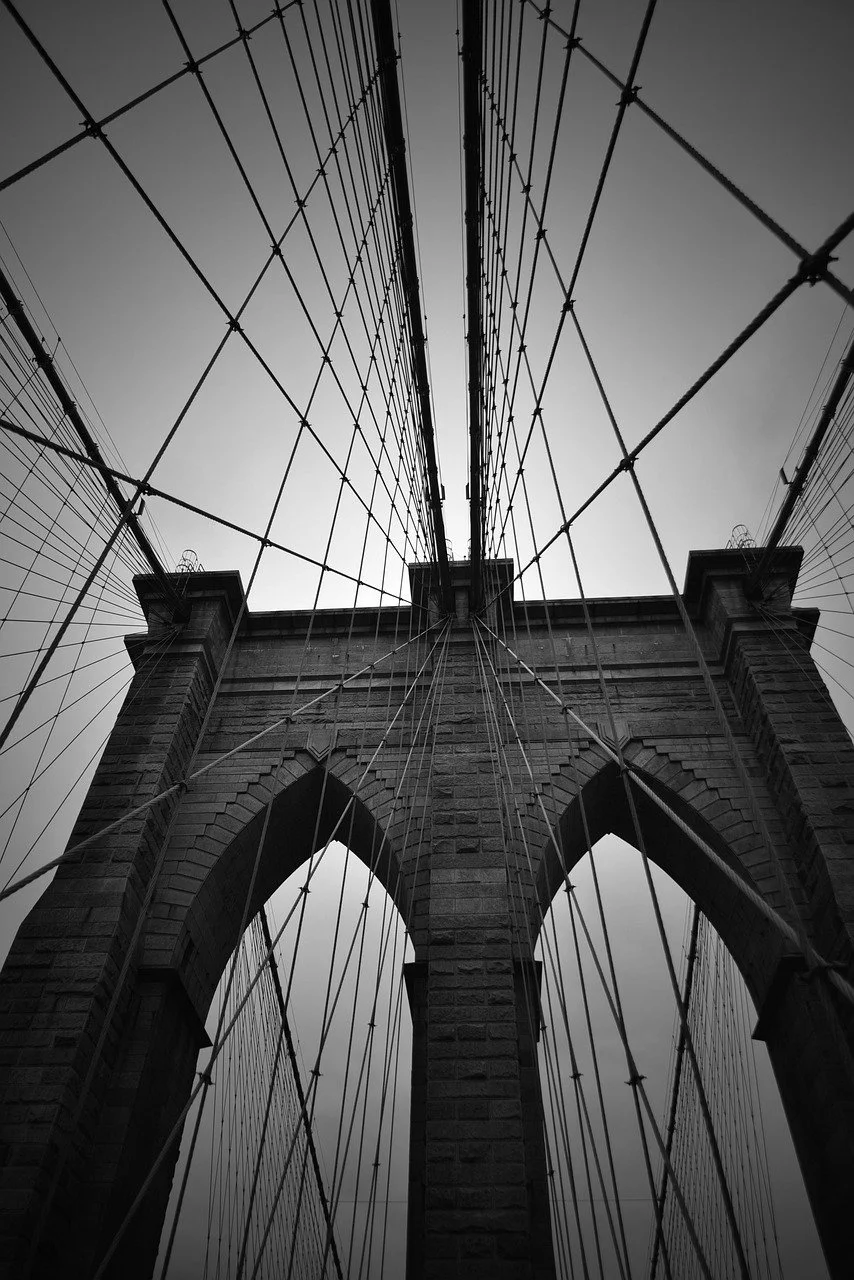 Black and white photo of the Brooklyn Bridge taken from below, showing stone towers and numerous suspension cables.