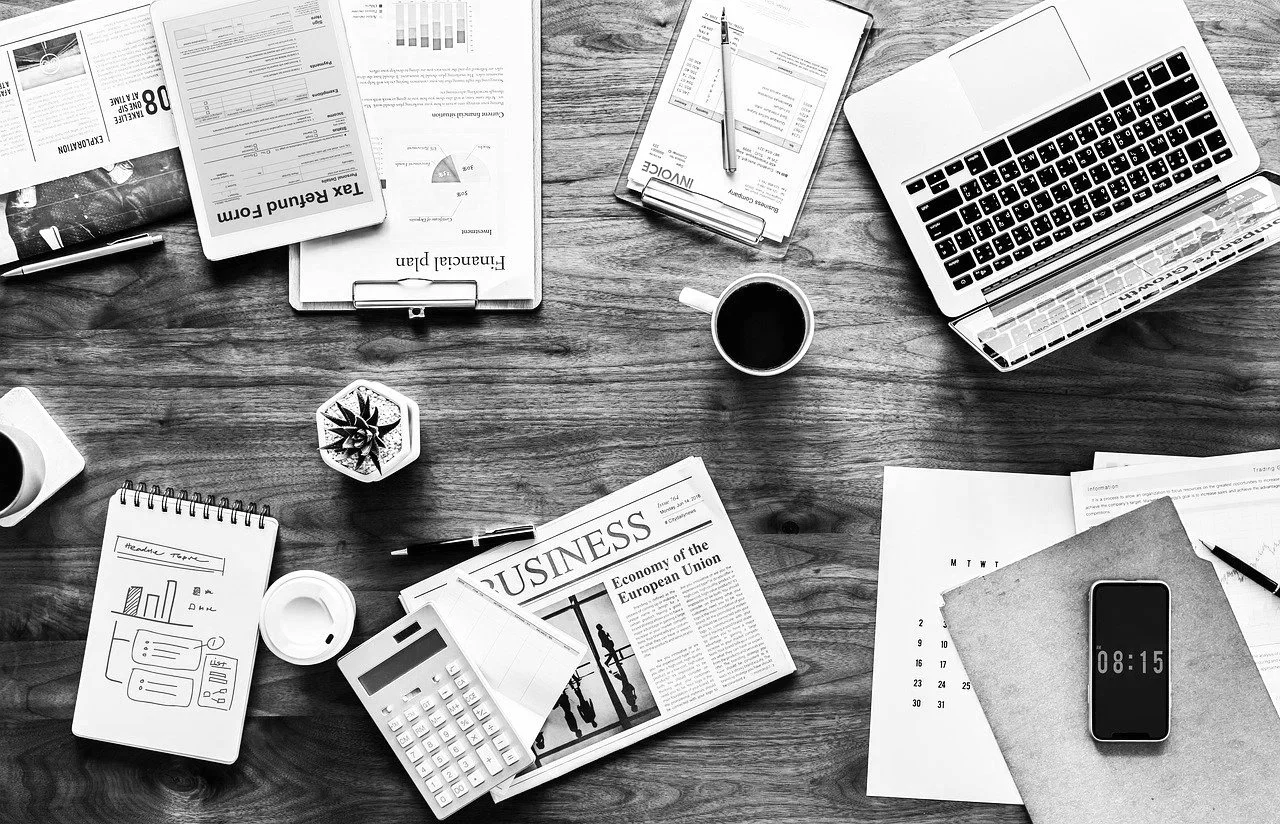 A workspace with financial documents, a laptop, a smartphone, a calendar, a notepad with charts, cups of coffee, and office supplies arranged on a wooden table.