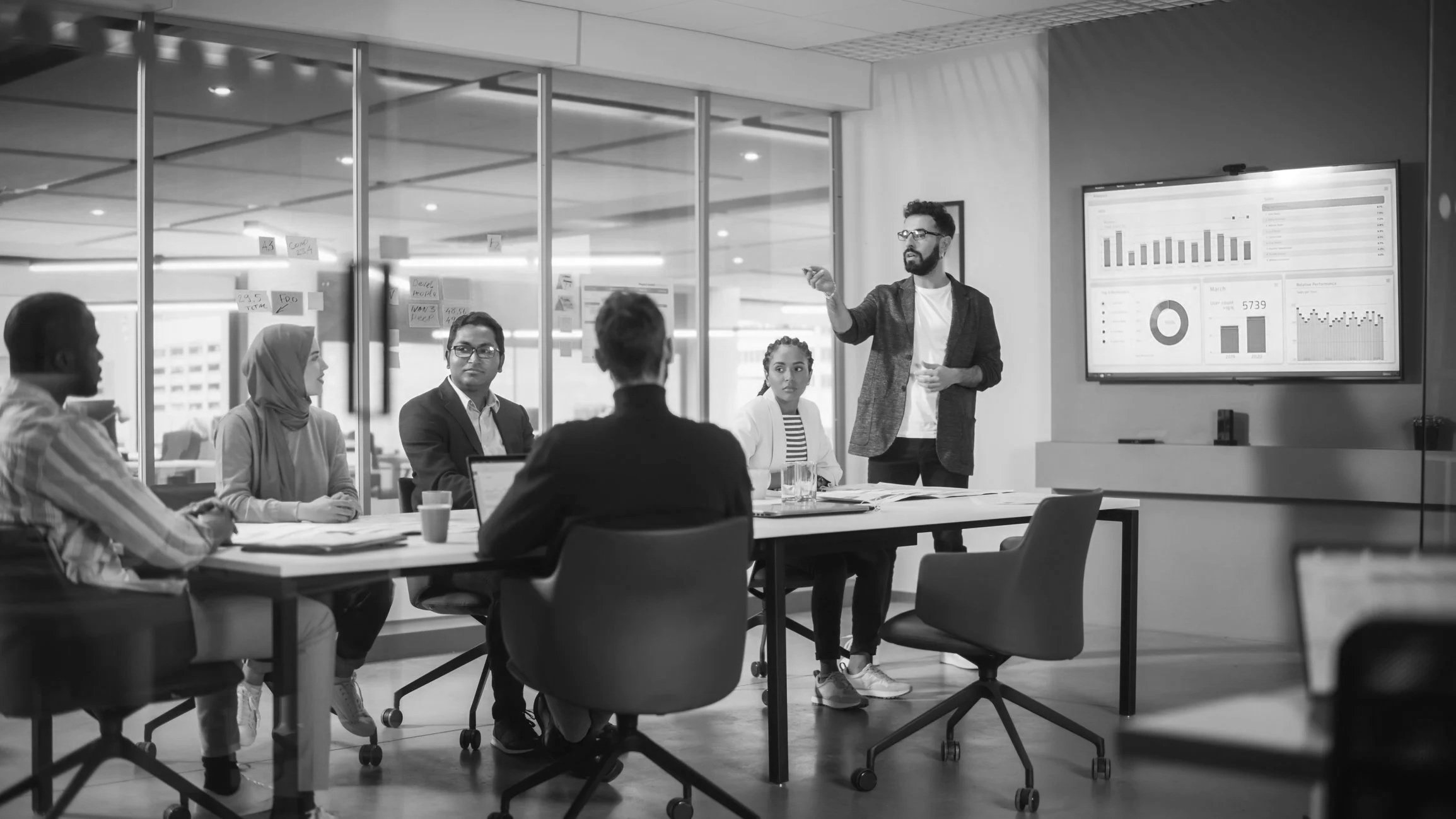 A group of five people in a business meeting room, seated around a table, listening to a man standing and presenting data on a large screen displaying graphs and charts.
