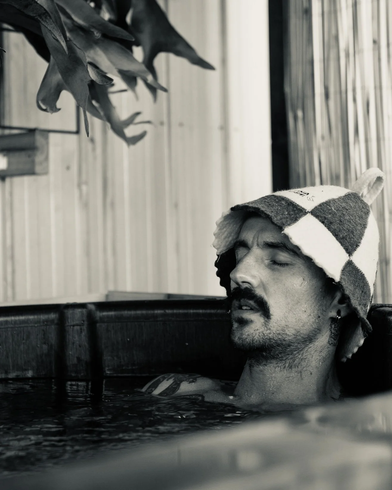 A man with a beard and closed eyes is relaxing in a wooden hot tub, wearing a checkered towel or hat on his head, with a wooden wall in the background.