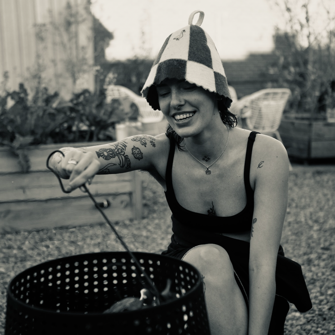 A smiling young woman with tattoos, wearing a checkered bucket hat and a tank top, reaching into a trash can in an outdoor setting.