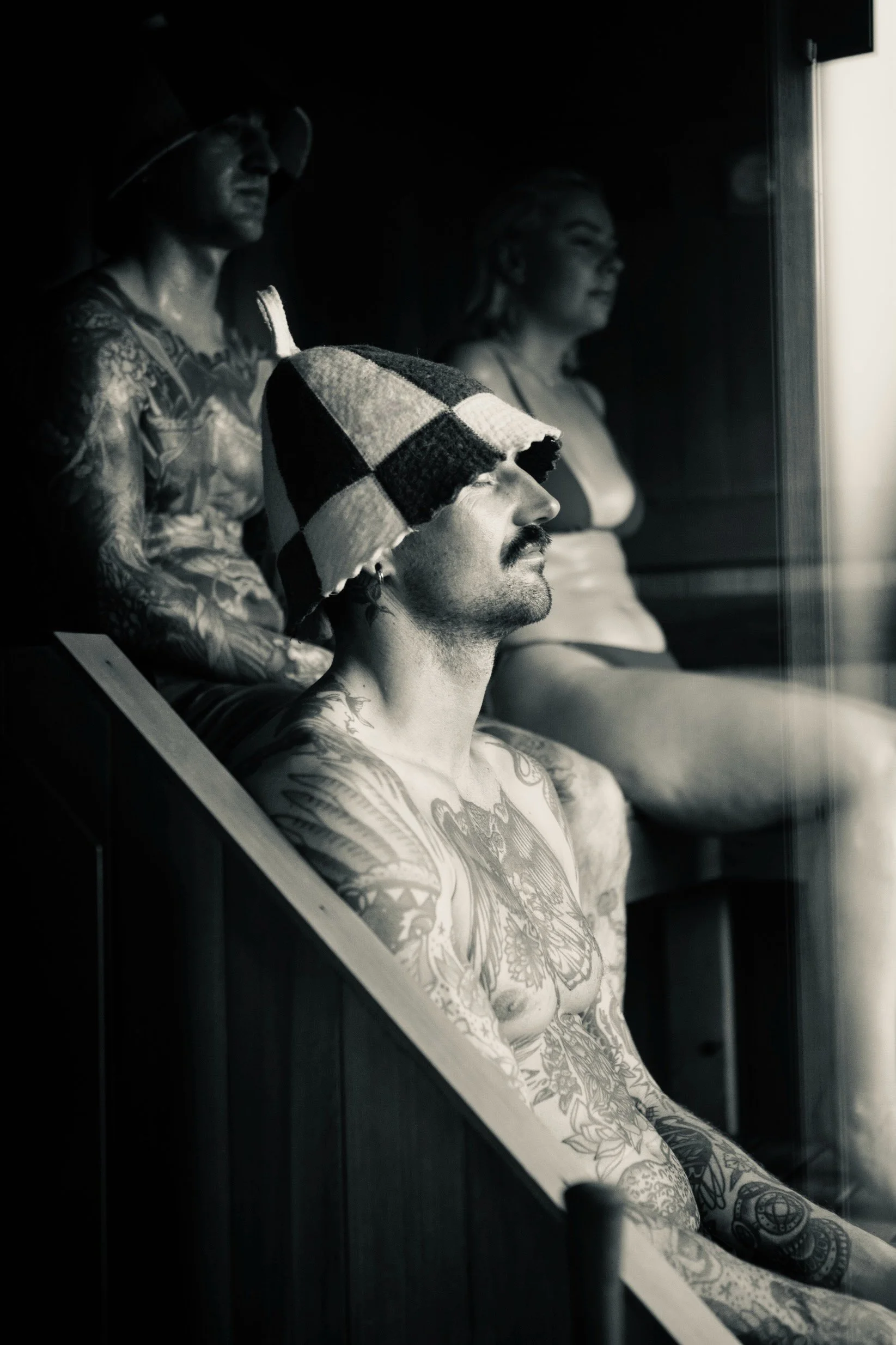 A black-and-white photo of three tattooed individuals sitting and looking out a window, with one person in the foreground wearing a checkered hat and sporting tattoos on his chest and arms.