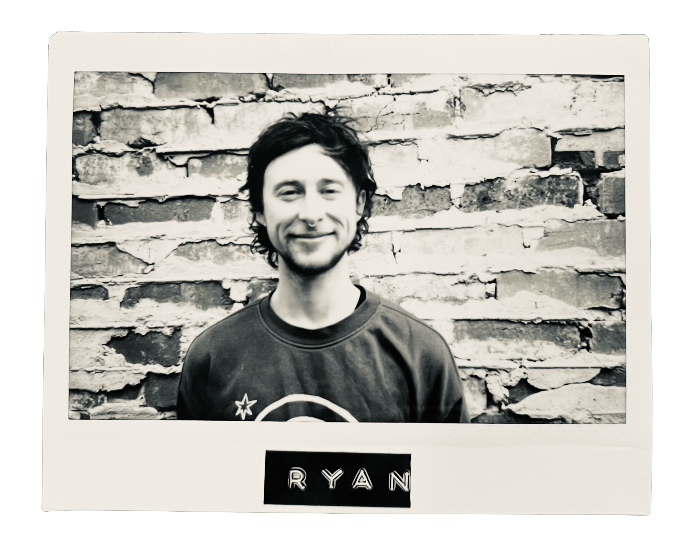 Black-and-white photograph of a smiling young man with curly hair standing in front of a brick wall, wearing a sweatshirt with a star and circular design, with the name blend labeled 'RYAN' at the bottom.