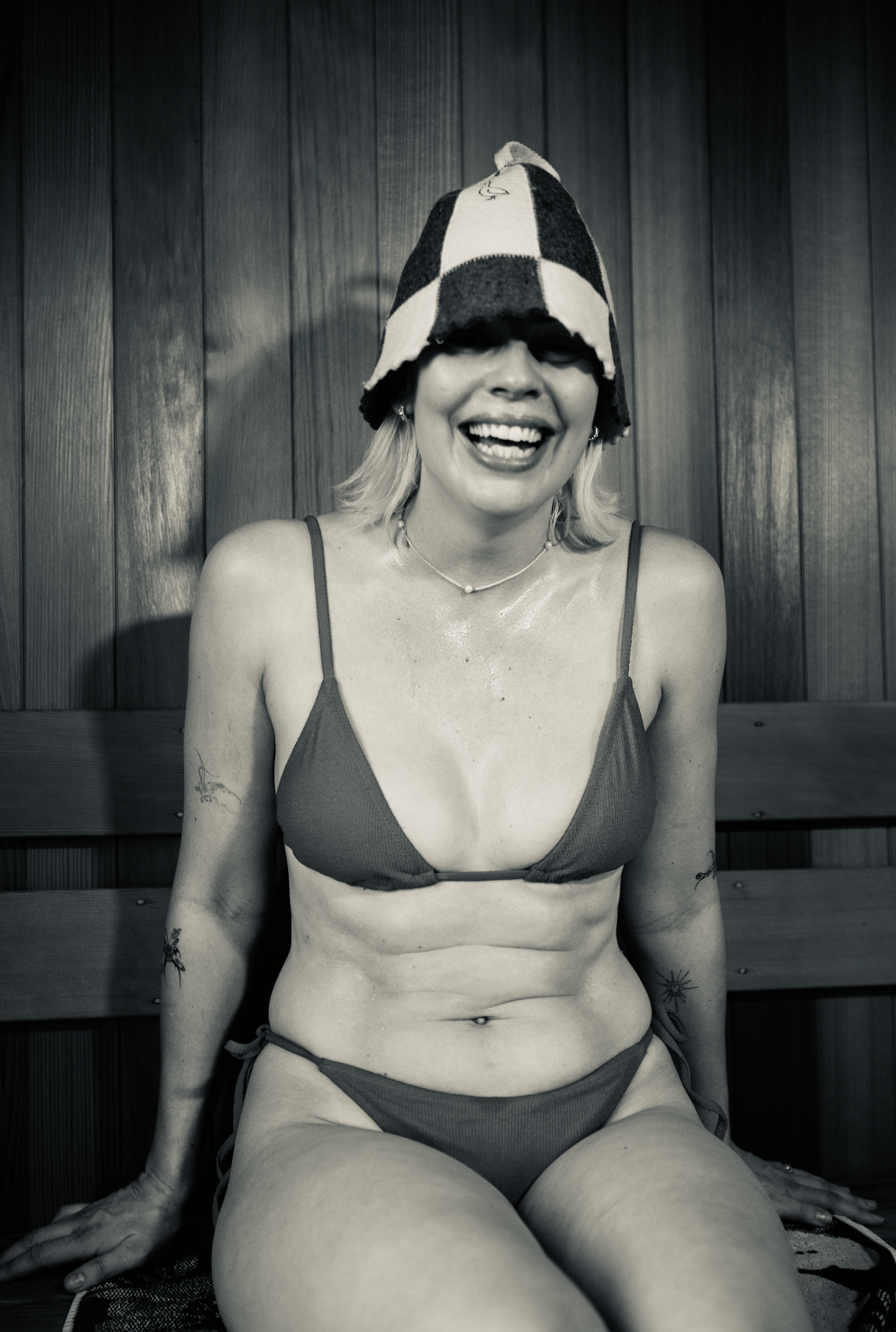 A smiling woman in a bikini and a checkered beanie sitting on a wooden bench against a wood-paneled wall.