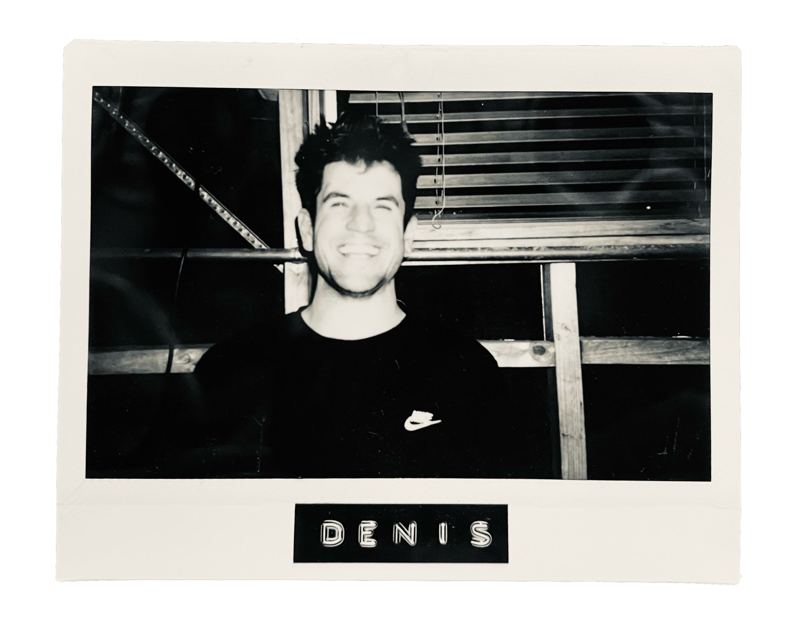 Black and white photo of a smiling man with dark, spiky hair, wearing a Nike shirt, standing against a background of wooden slats and a pipe, with the name "Dennis" on the bottom label.