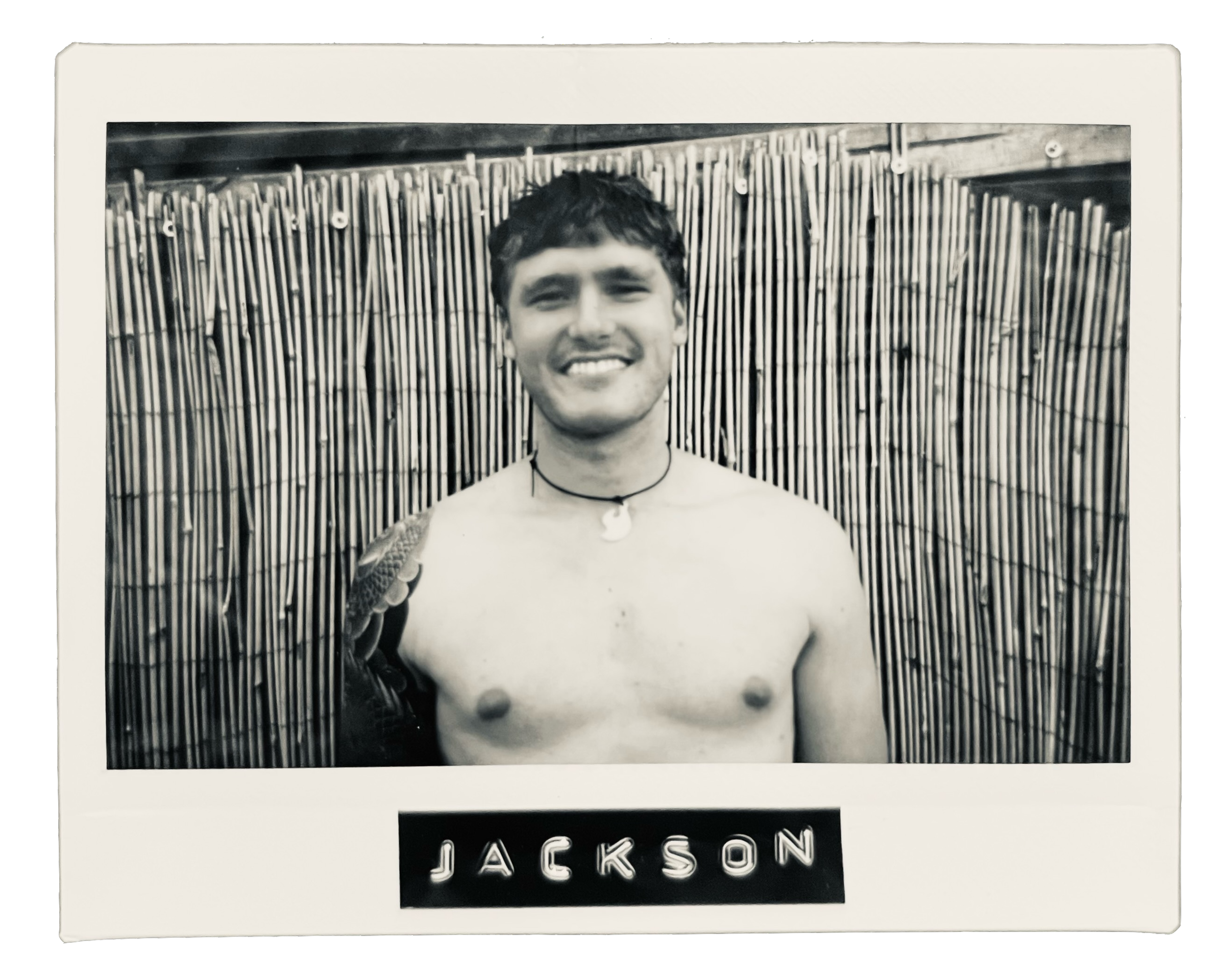 Black and white photograph of a smiling young man with dark hair, no shirt, wearing a necklace, standing in front of a bamboo fence. The photo has a caption reading 'JACKSON'.