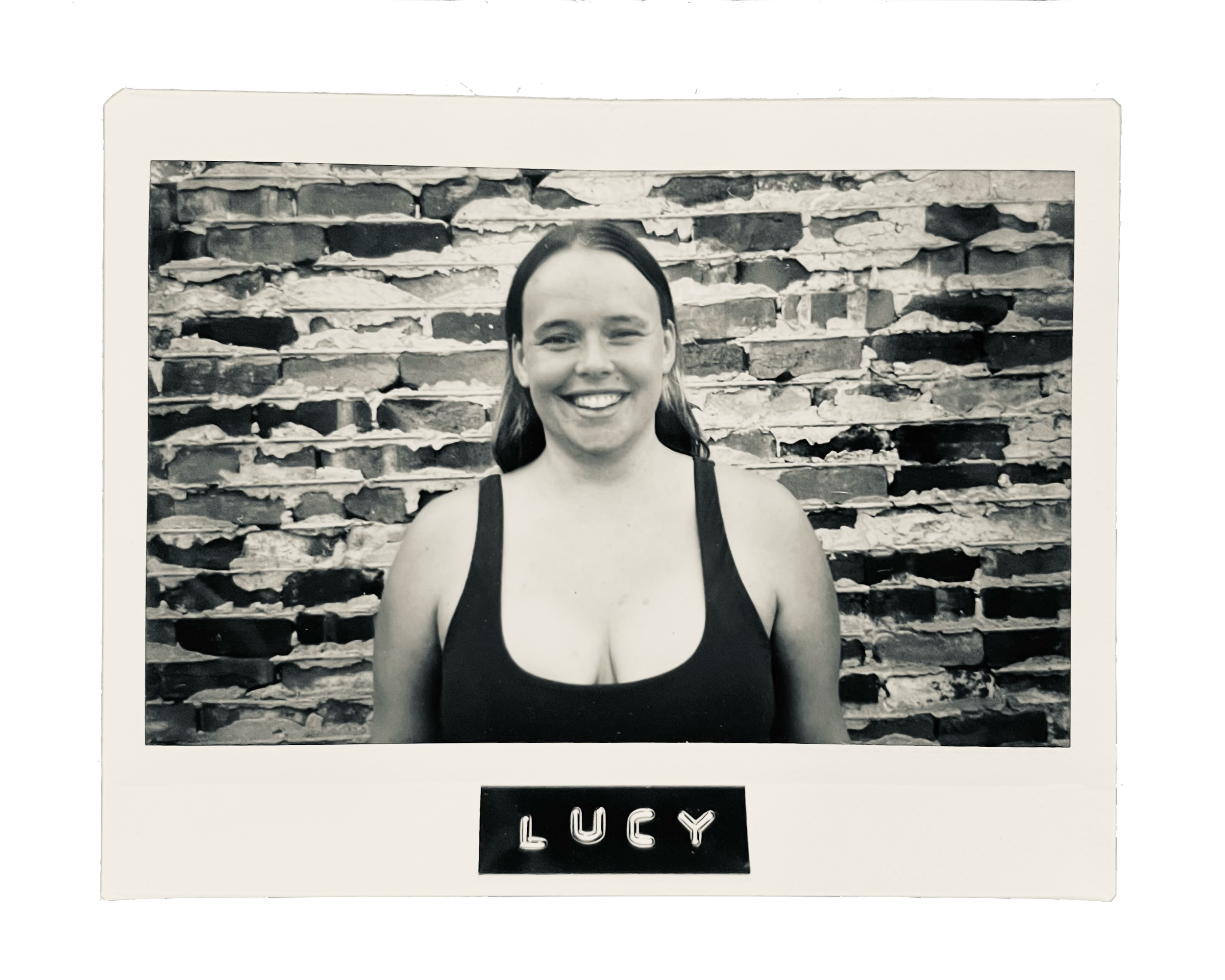 A black and white photograph of a smiling woman with shoulder-length hair wearing a sleeveless top, standing in front of a textured brick wall. The photo is framed like an instant photograph with the name 'LUCY' written on a label at the bottom.