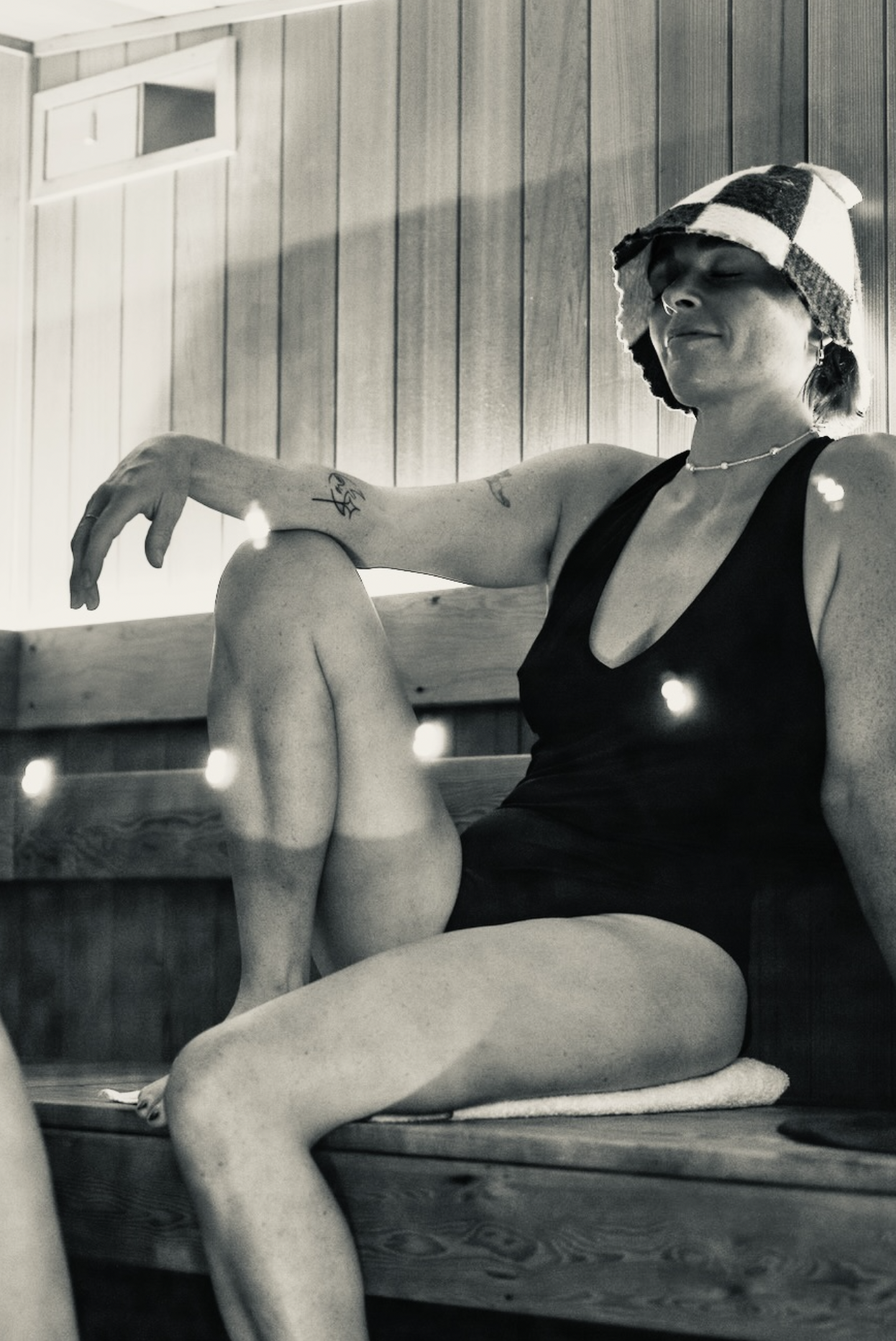 A woman sitting on a wooden bench with her legs crossed, wearing a sleeveless black top and a checkered hat, with her eyes closed and a peaceful expression.