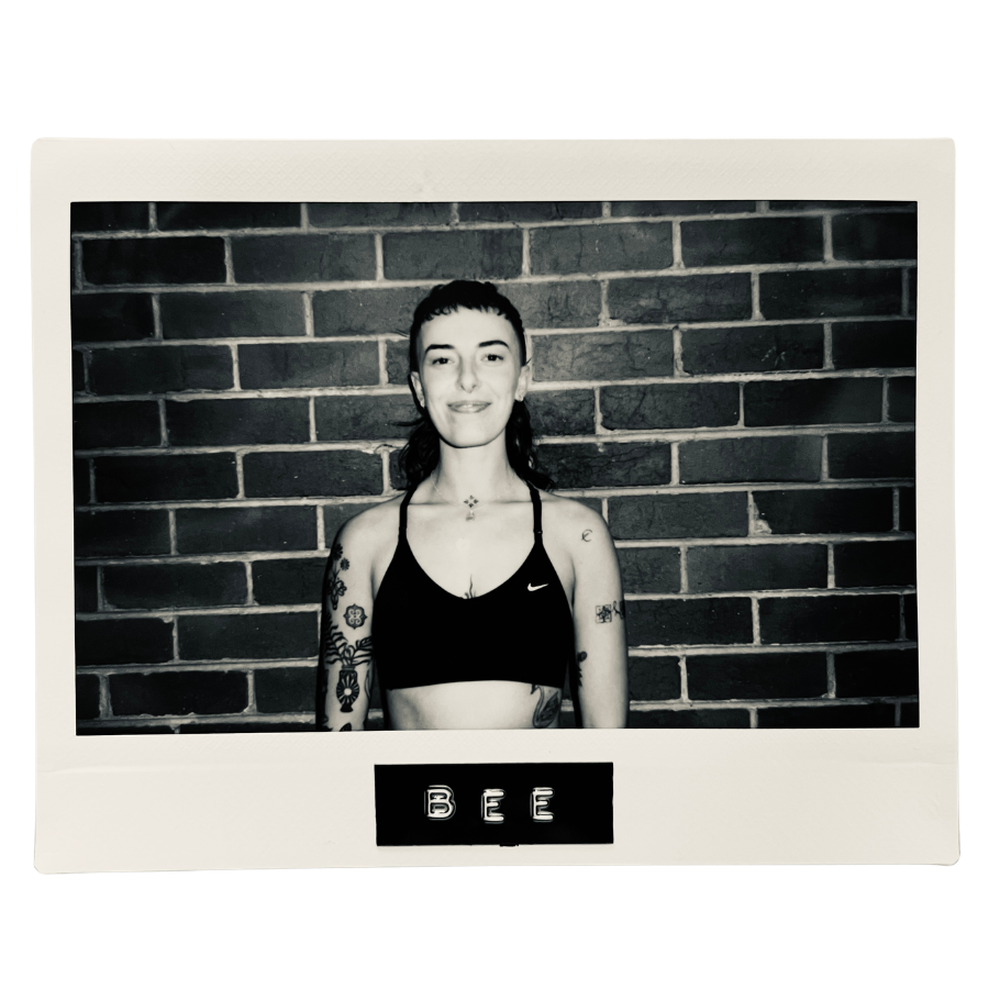 Black and white photograph of a woman with tattoos wearing a sports bra, standing in front of a brick wall, smiling at the camera.