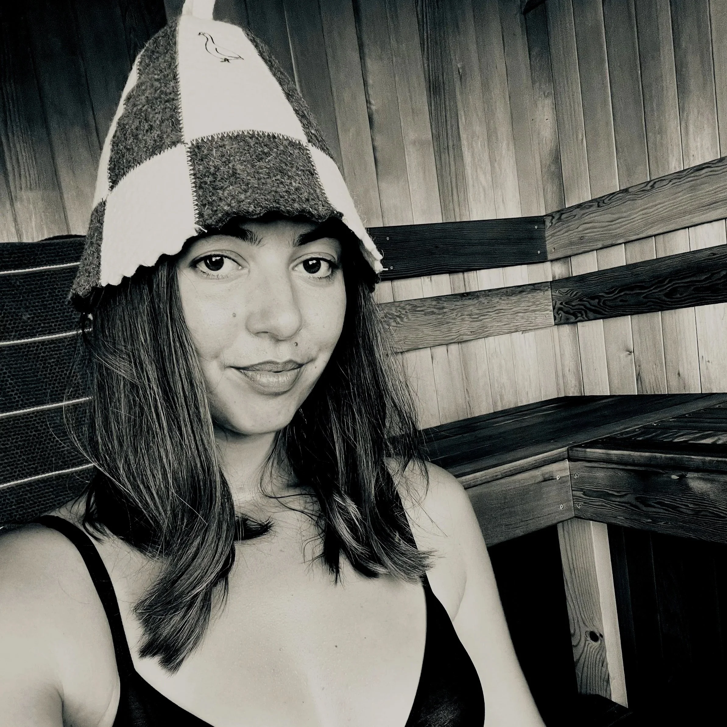 A woman with long dark hair, wearing a checkered hat, smiling slightly, in a setting with wood-paneled walls.